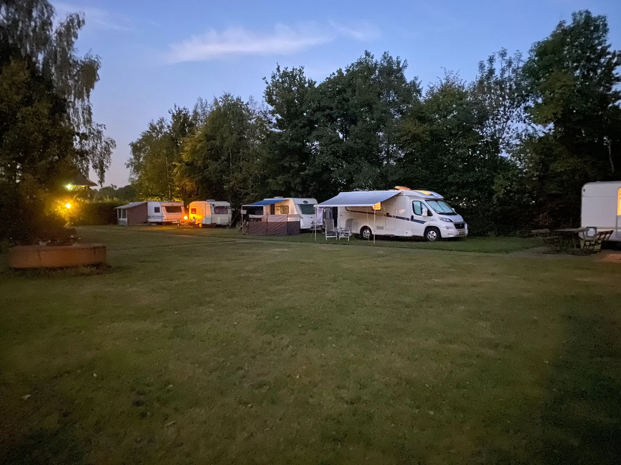 Een kampeerplek met meerdere campers en caravans op een grasveld, omgeven door bomen, tijdens de schemering of avond.