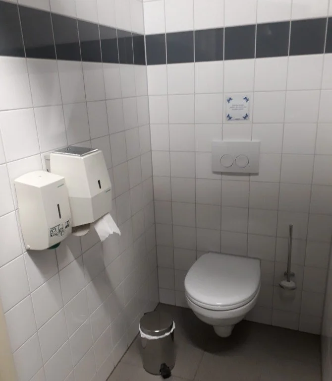 Kleine toiletruimte met witte tegels, wandtoiletten, een bidetborstel, een afvalbak met deksel en handdoekdispensers.