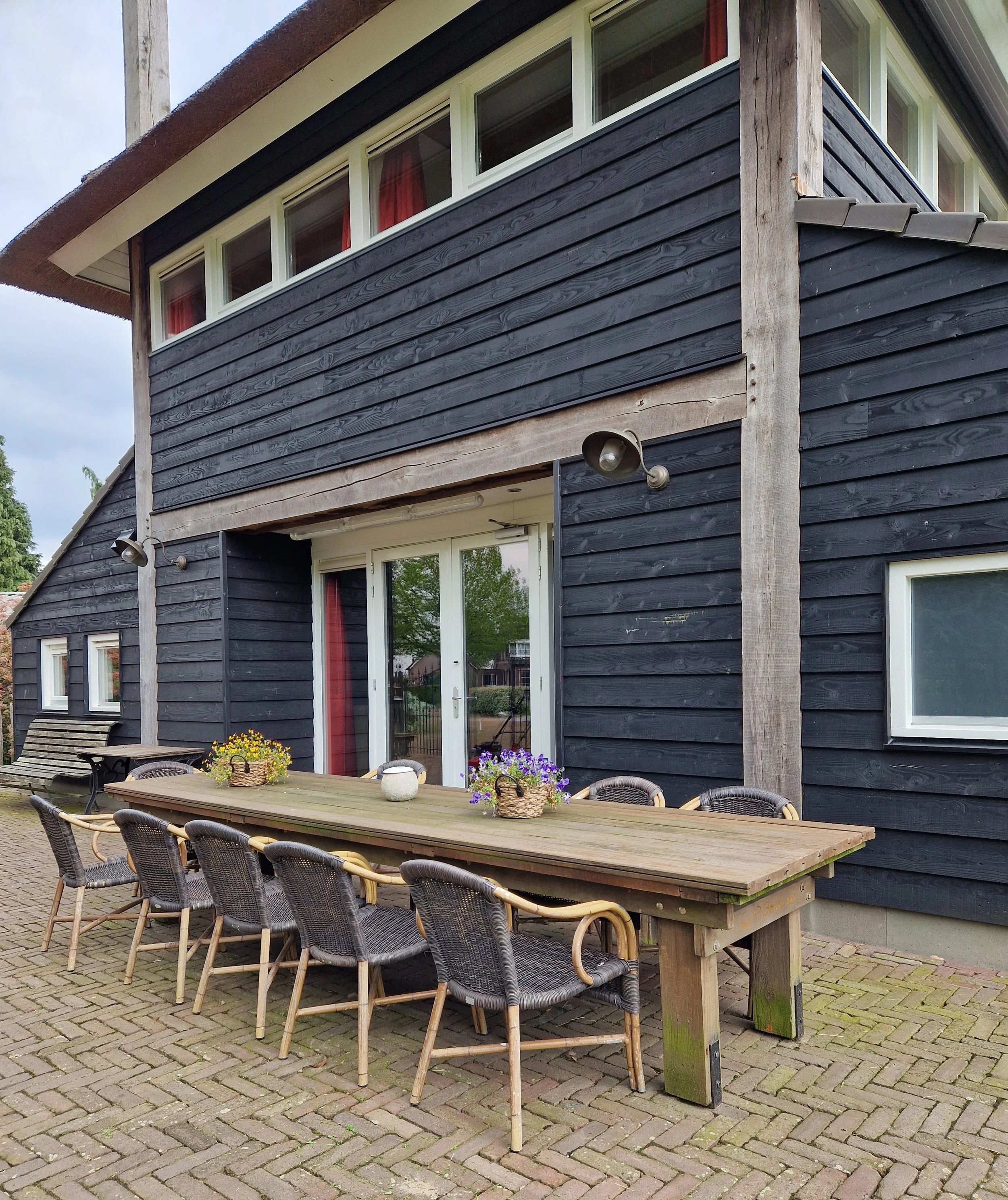Uitzicht van een buitenzet met een houten tafel en zes rieten stoelen voor een donkerhouten huis met meerdere ramen en een glazen schuifdeur, omgeven door een betegelde patio.