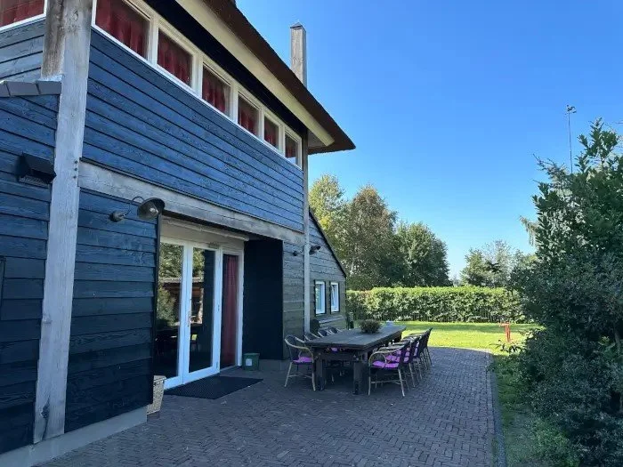 Buitenpatio met tuintafel en stoelen naast een modern huis met grote ramen en blauwe houten buitenbekleding, omgeven door groene planten en bomen onder een heldere blauwe lucht.