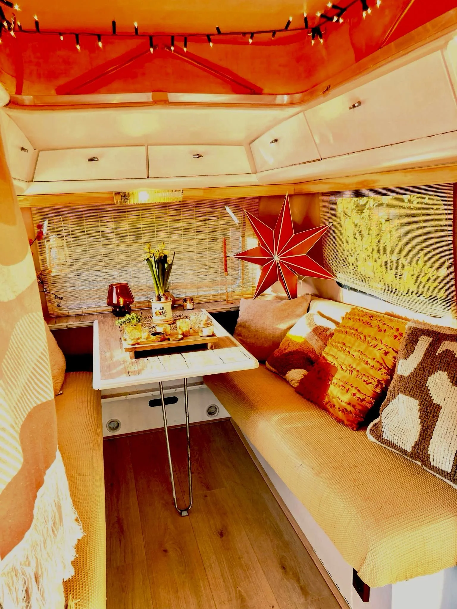 Een knusse camper interieur met een gele bank, een tafel met decoraties, een rode sterrenlamp en vensters met bamboe jaloezieën, alles in warme kleuren.