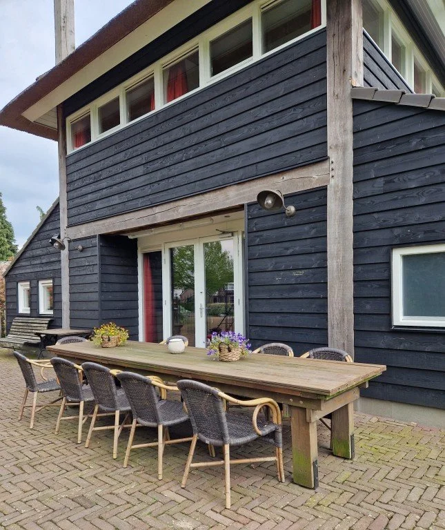 Een buiten tafellounge met negen stoelen voor een huis met donkere houten gevelbekleding en grote ramen.