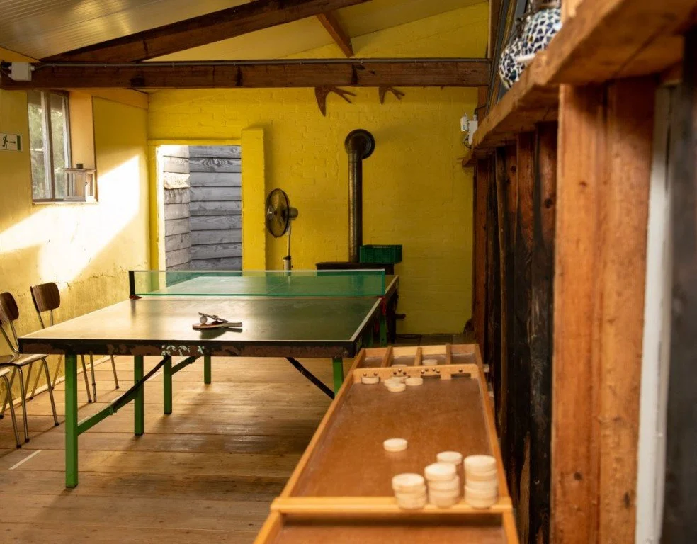 Een recreatieruimte met een tafeltennistafel, een ventilator, een schoorsteen, en een houten plankenkast. Er zijn drie zwarte stoelen langs de muur en een raam dat het binnen verlicht.