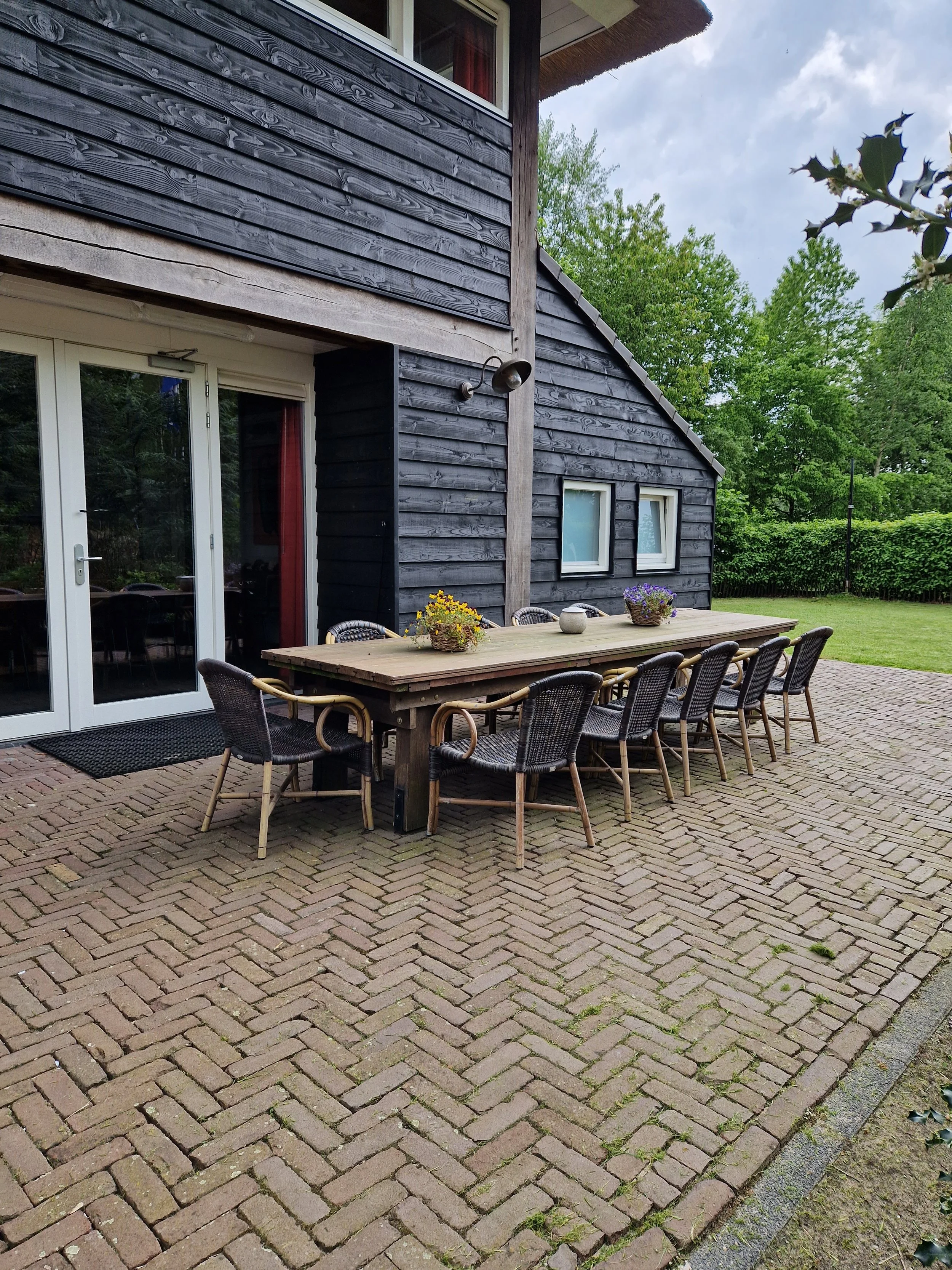 Een buiten eethoek met een lange houten tafel en acht rieten en houten stoelen op een bakstenen patio naast een donker houten huis met twee kleine ramen en een glazen deur.