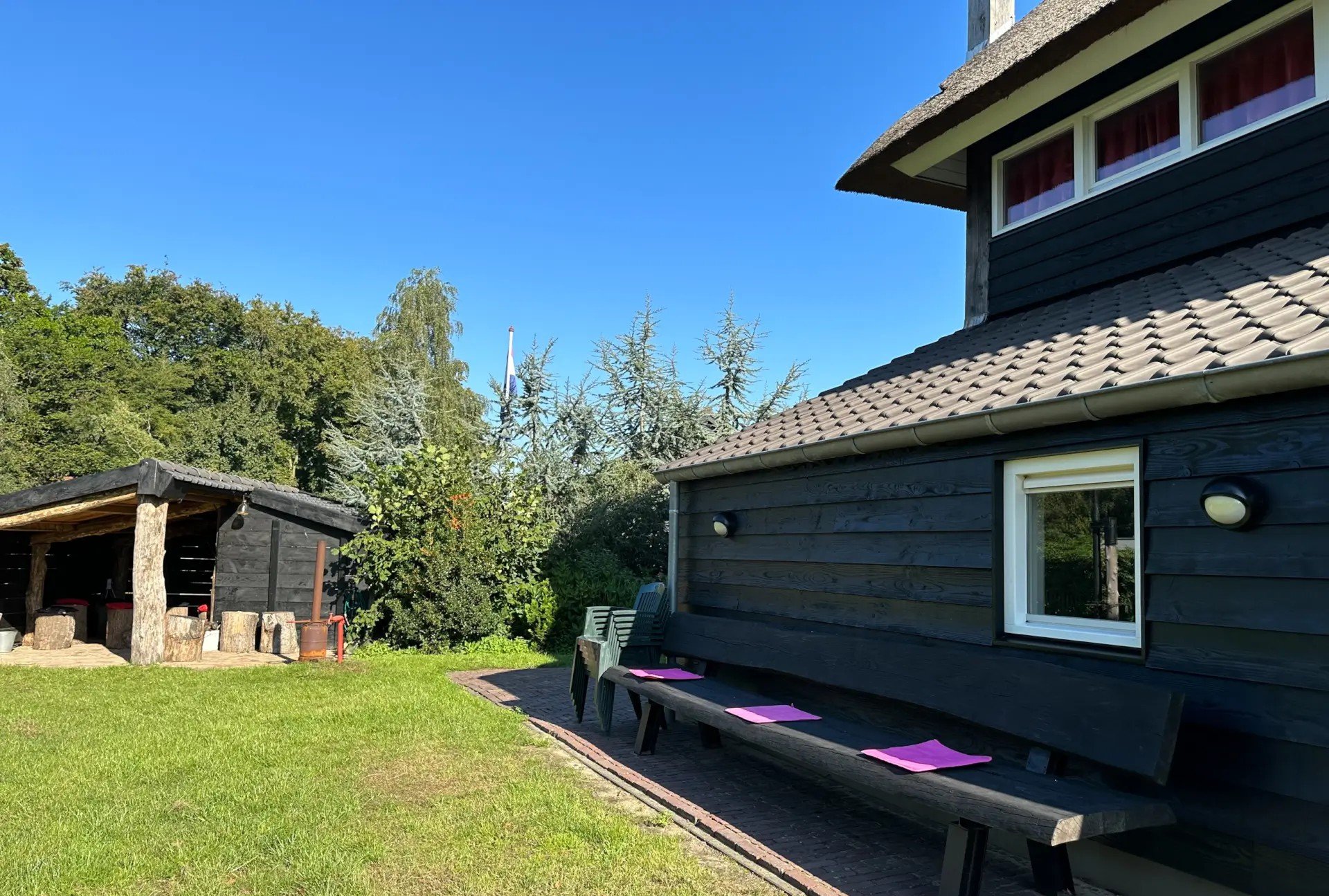 Buitenruimte met een donker houten huis, een grasveld, een houten bank met drie paarse placemats, en een schaduwrijke houten overkapping op een zonnige dag.