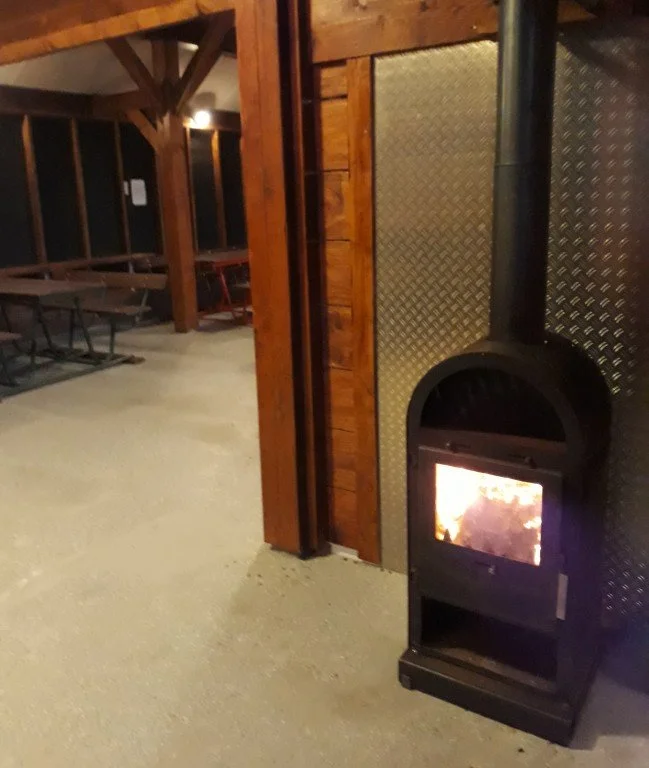 Binnenkant van een houten en metalen gebouw met een houtkachel die brandt.