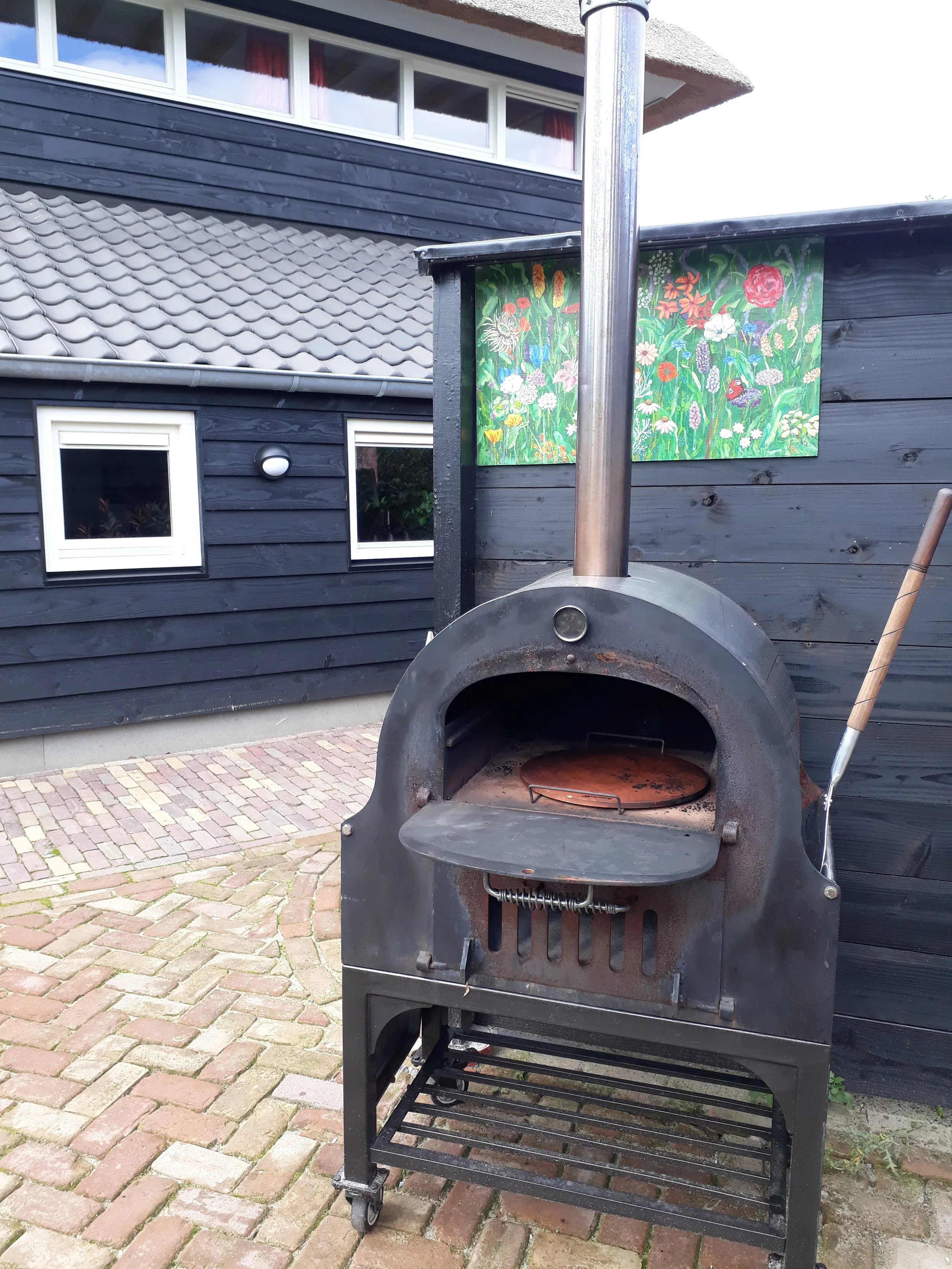 Een buitenoven met een metalen deksel en een roestig uiterlijk staan op een grindpad voor een zwarte houten gevel. Op de gevel hangen twee kleurrijke schilderijen met bloemen.