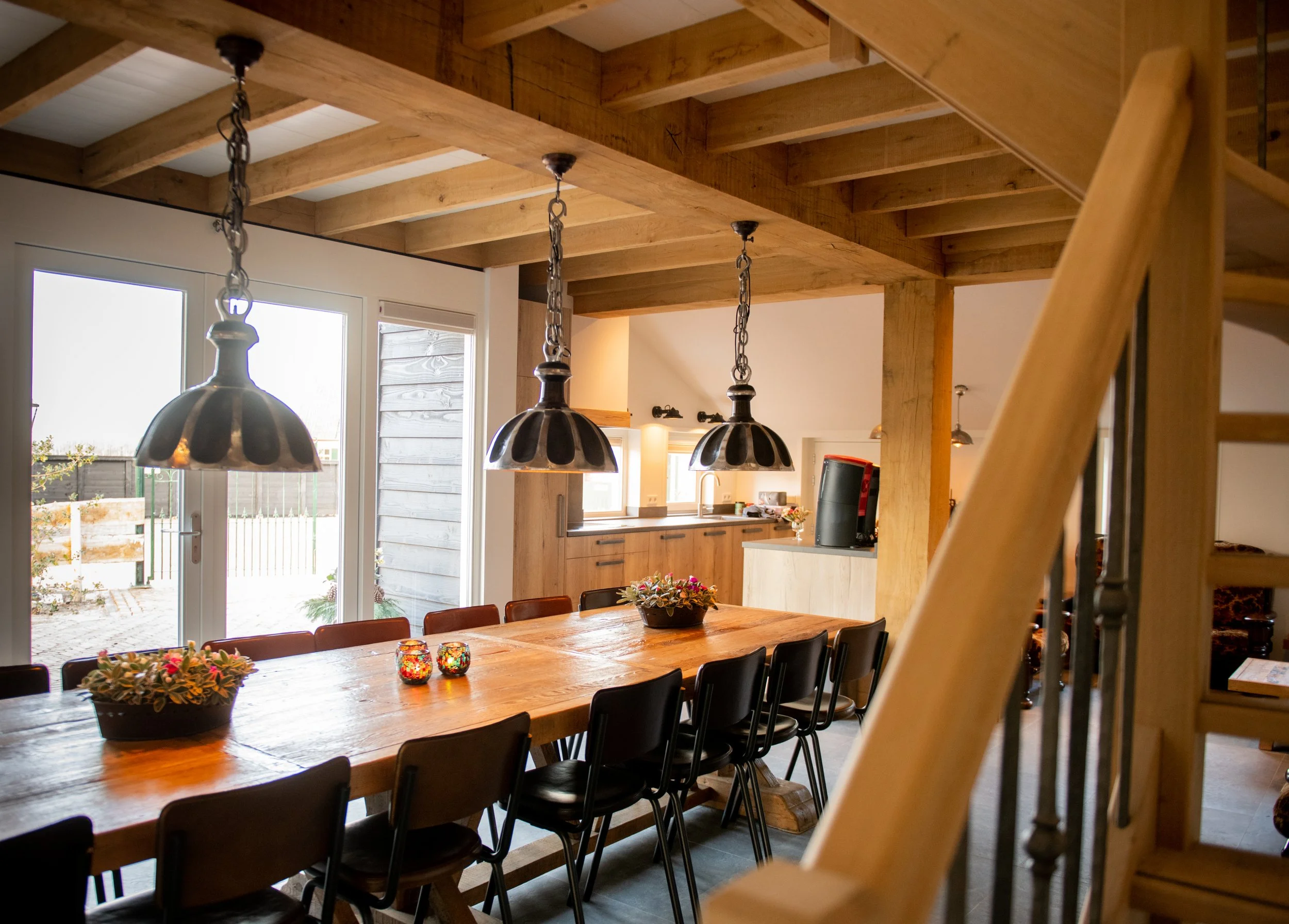 Binnenkijker van een eetkamer met een grote houten tafel, zwarte stoelen, hanglampen, grote ramen en een keuken in de achtergrond.