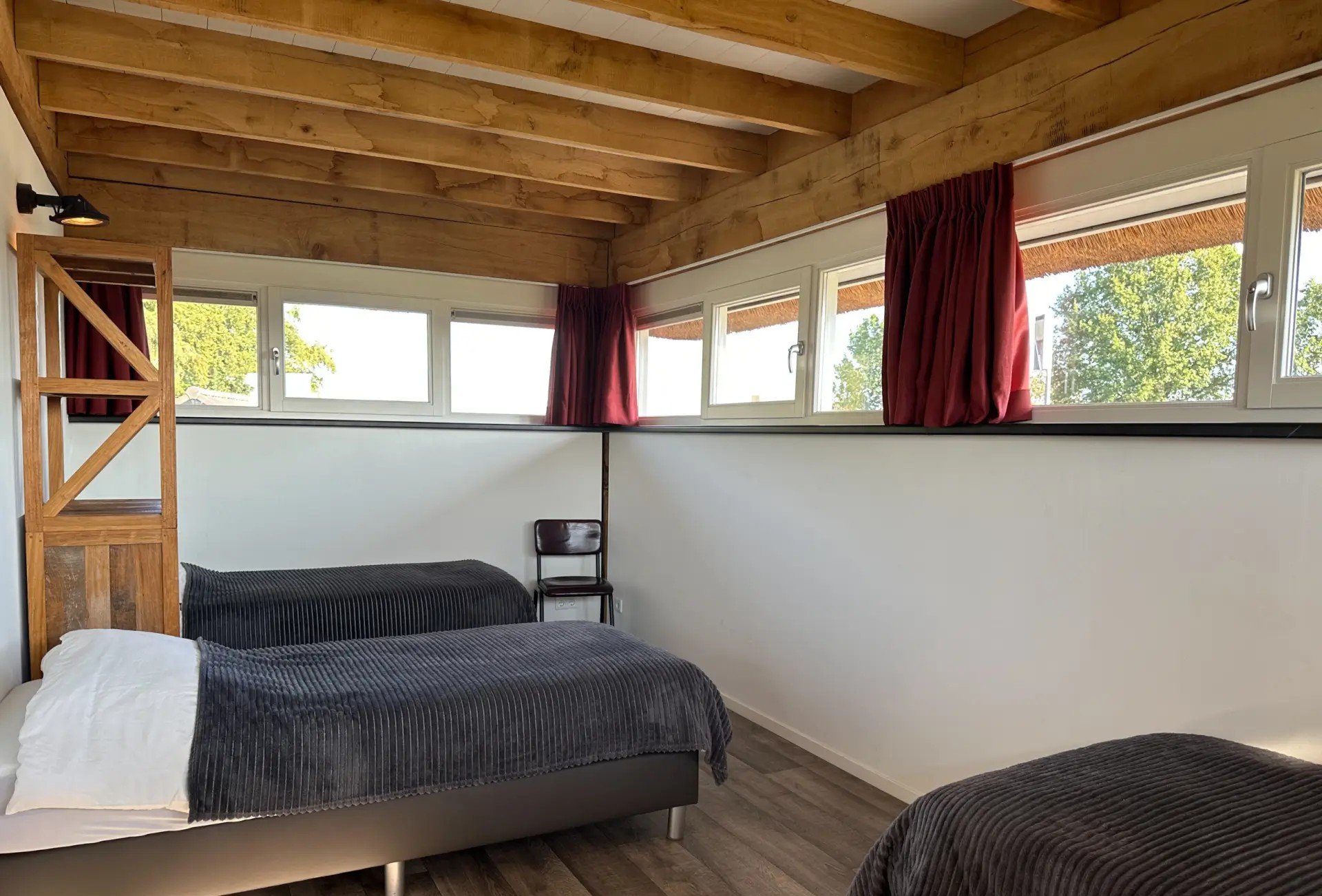 Een kamer met twee bedden met donkergrijze dekens, een houten plafond en een lange rij ramen met rode gordijnen, uitzicht op groene bomen buiten.