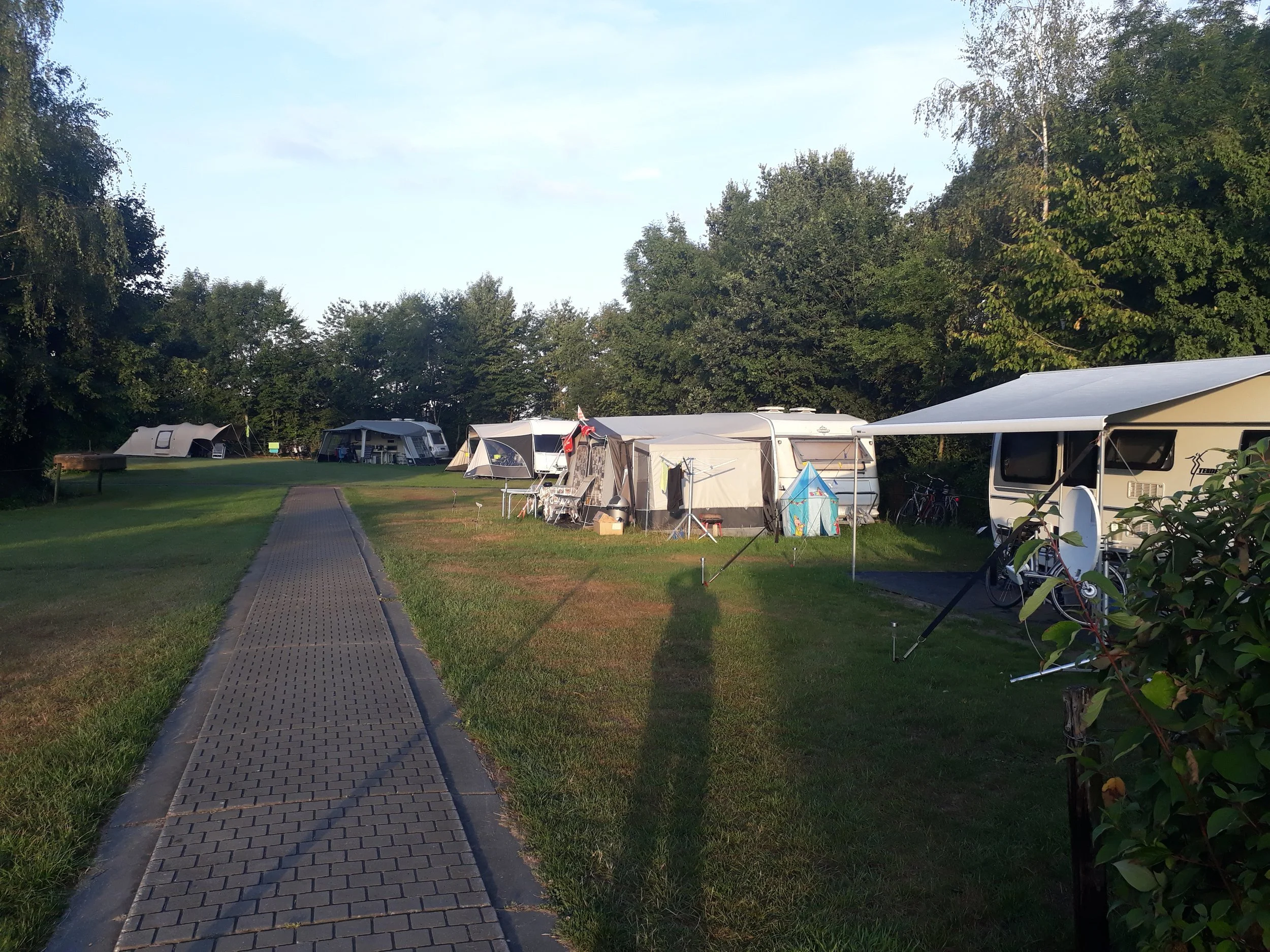 Beekpark met campingplaatsen, caravans, en campingartikelen, omringd door groene bomen en gras op een zonnige dag.