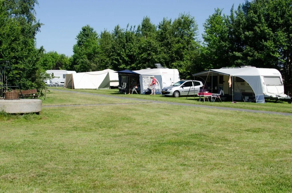 Campingplaats met caravans, tenten en campers, gelegen op een grasveld met bomen op de achtergrond.