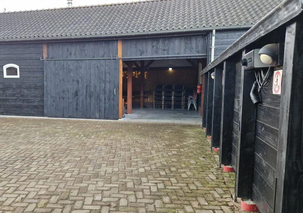 Een donkere schuur met een open dubbele deur, binnenin stapels stoelen en een brandblusser aan de muur.