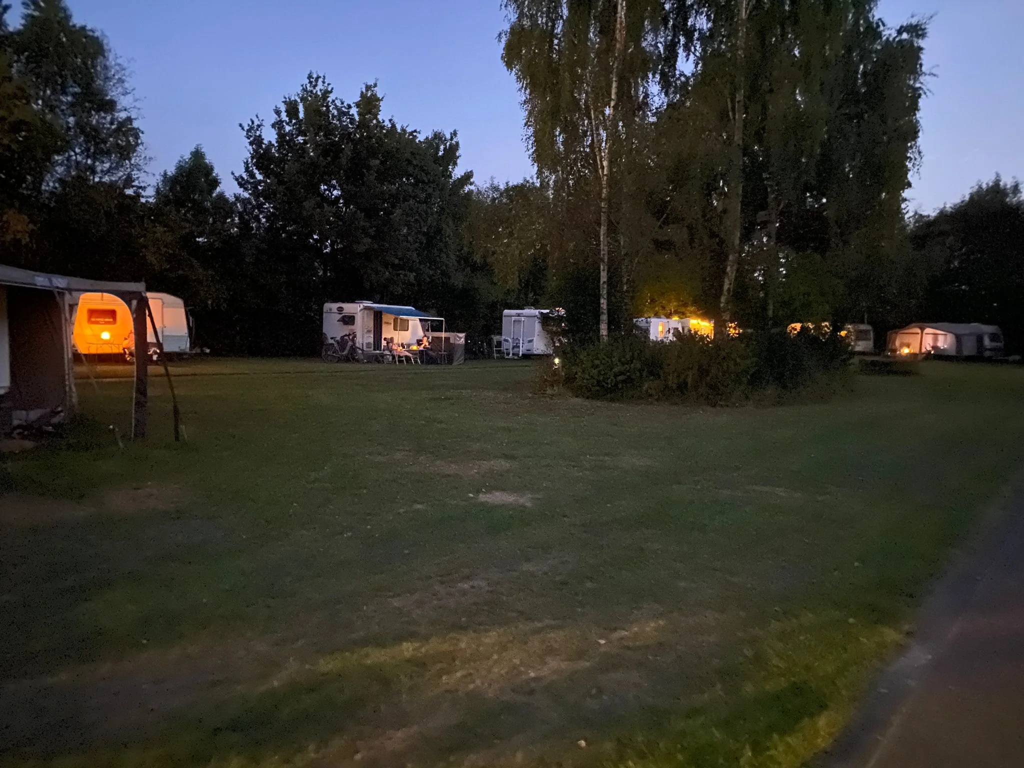 Campingveld met caravans en campers, verlichte tenten en bomen, 's avonds of nacht, rustige sfeer.