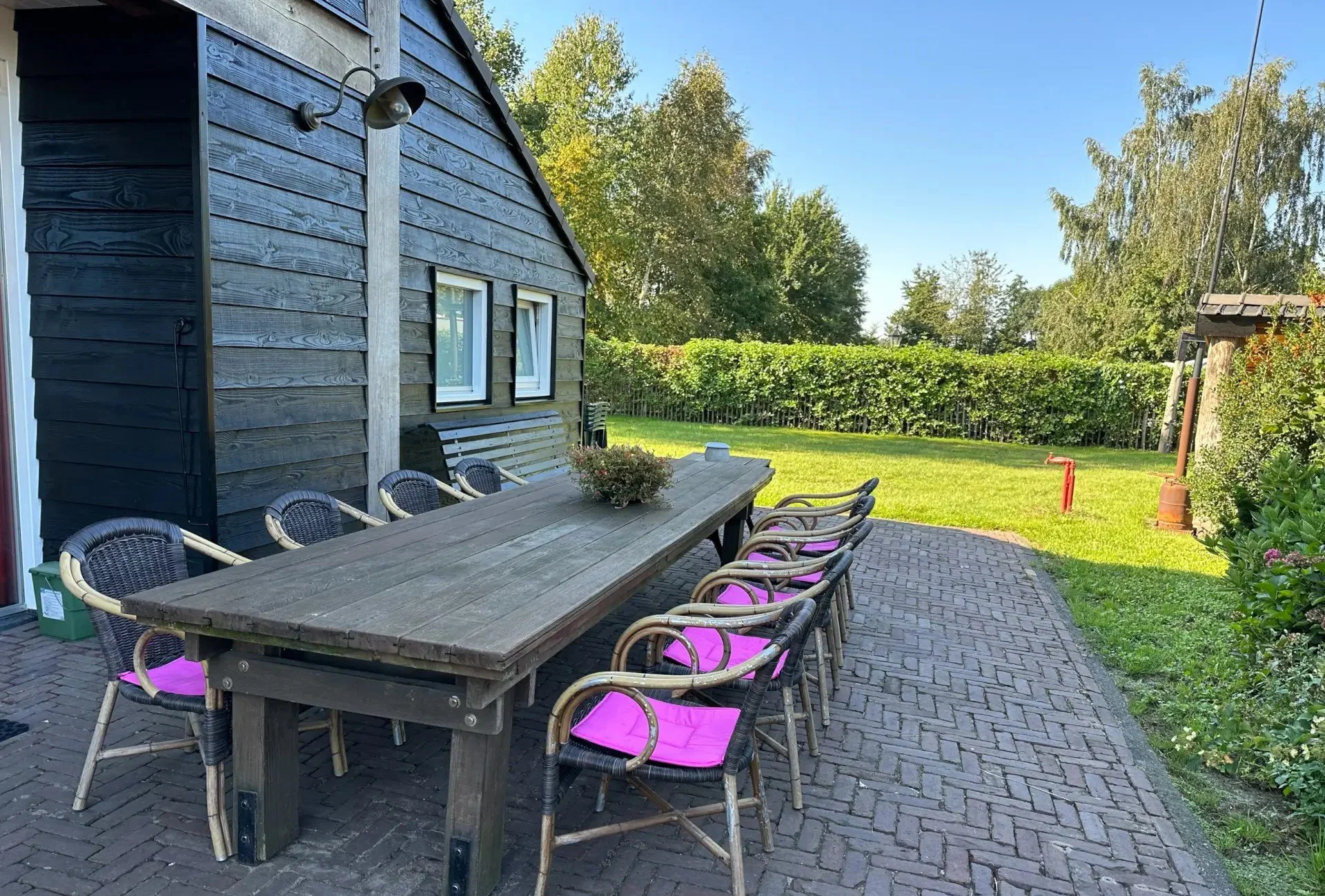 Buiten eetgedeelte met een lange houten tafel, acht wicker stoelen met roze zitkussens, naast een zwarte houten schuur en een groene tuin met bomen en een hek.