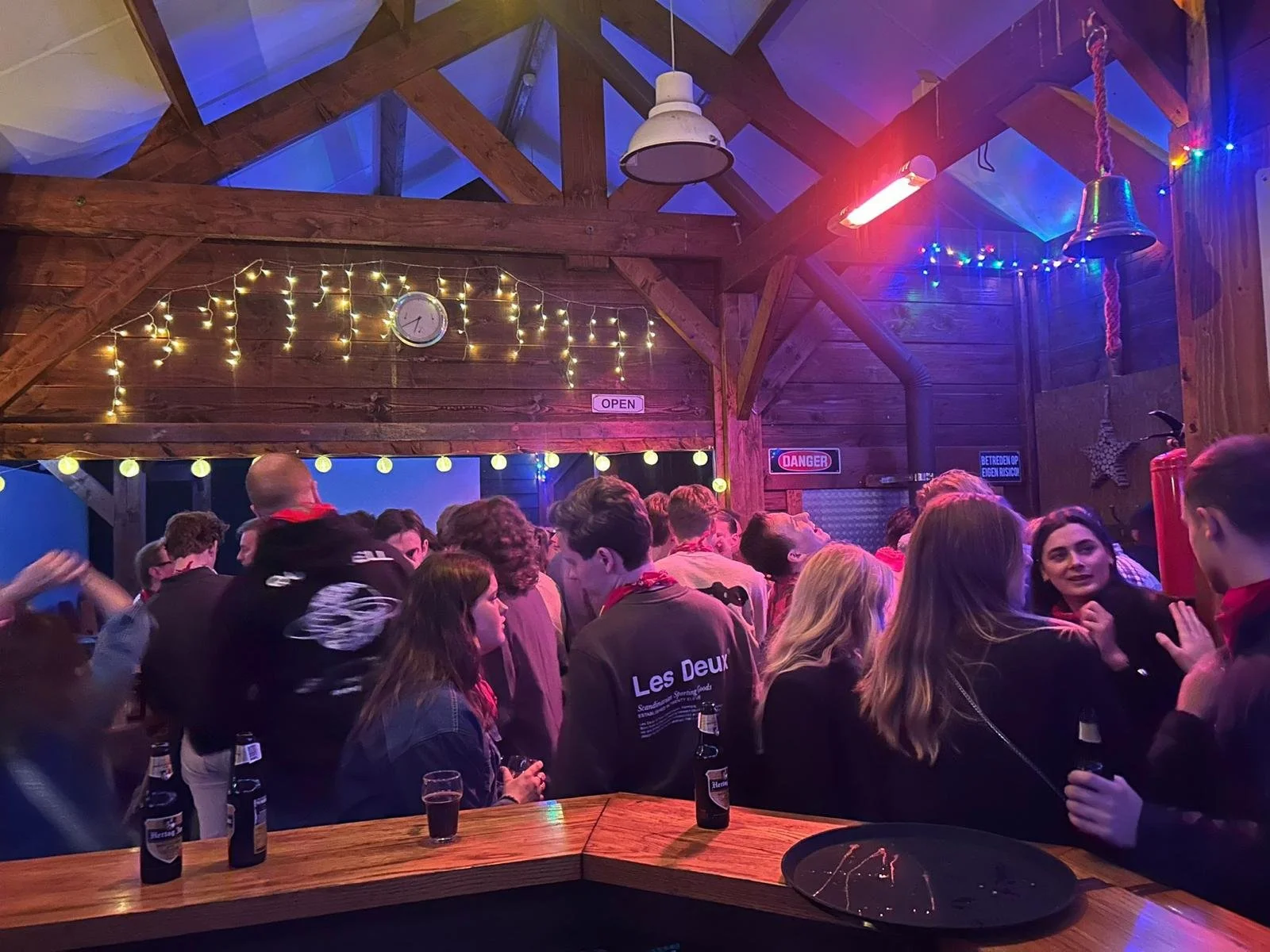 Mensen die binnen in een brouwerij of bar staan, met knusse houten muren en decoraties, waaronder lampjes, een klok en sterren. Er zijn verschillende mensen die praten en genieten, met bierflesjes op de bar.