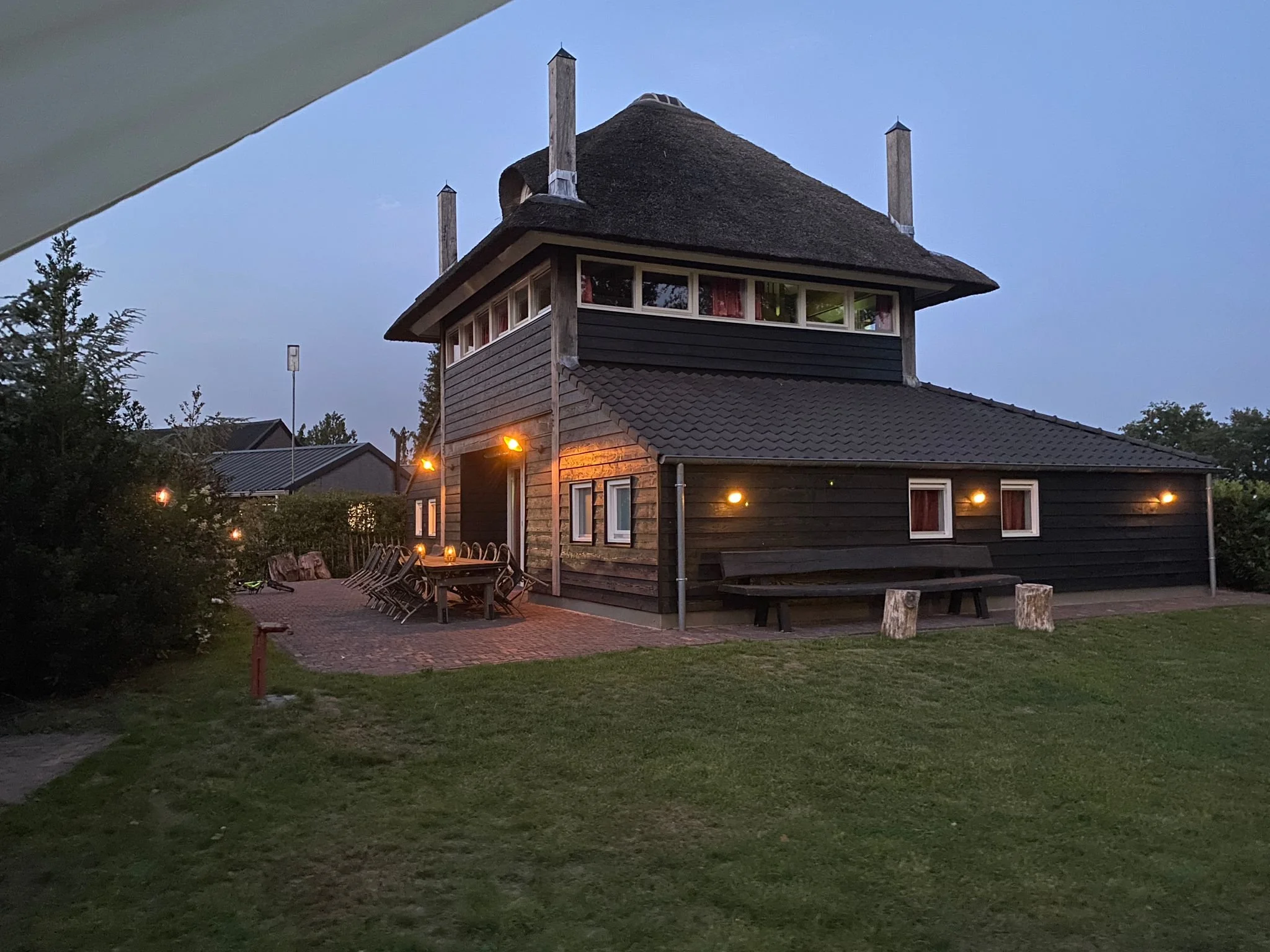 Een donkerkleurig, boerderijl soort huis met dat dak en een omringde tuin. Buiten staan er tafels en stoelen, en de tuin is verlicht met buitenspiegellampen, terwijl het schemer is.