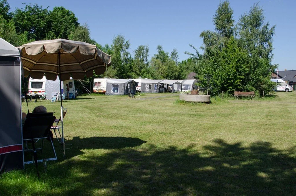 Een open grasveld met tenten en caravans, bomen op de achtergrond, en een persoon die onder een parasol zit in een camping of park.