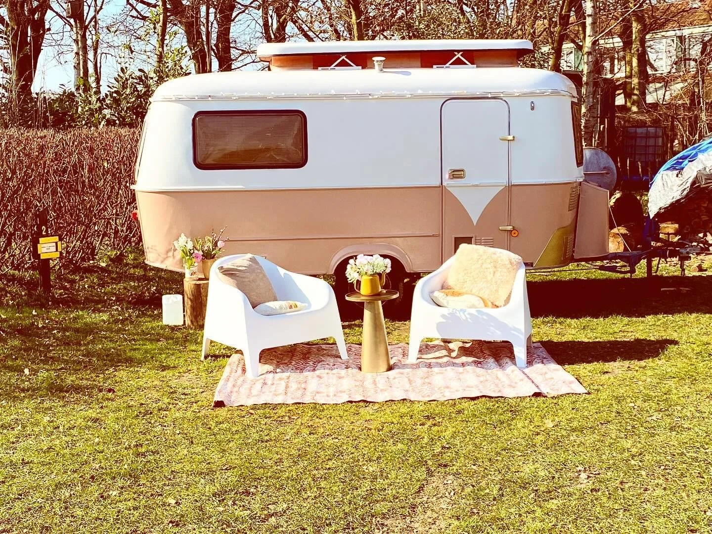 Twee witte stoelen met kussens, een kleine tafel met een bloemenvaas, gedrapeerd op een tapijt, voor een caravan op een grasveld, omgeven door bomen.