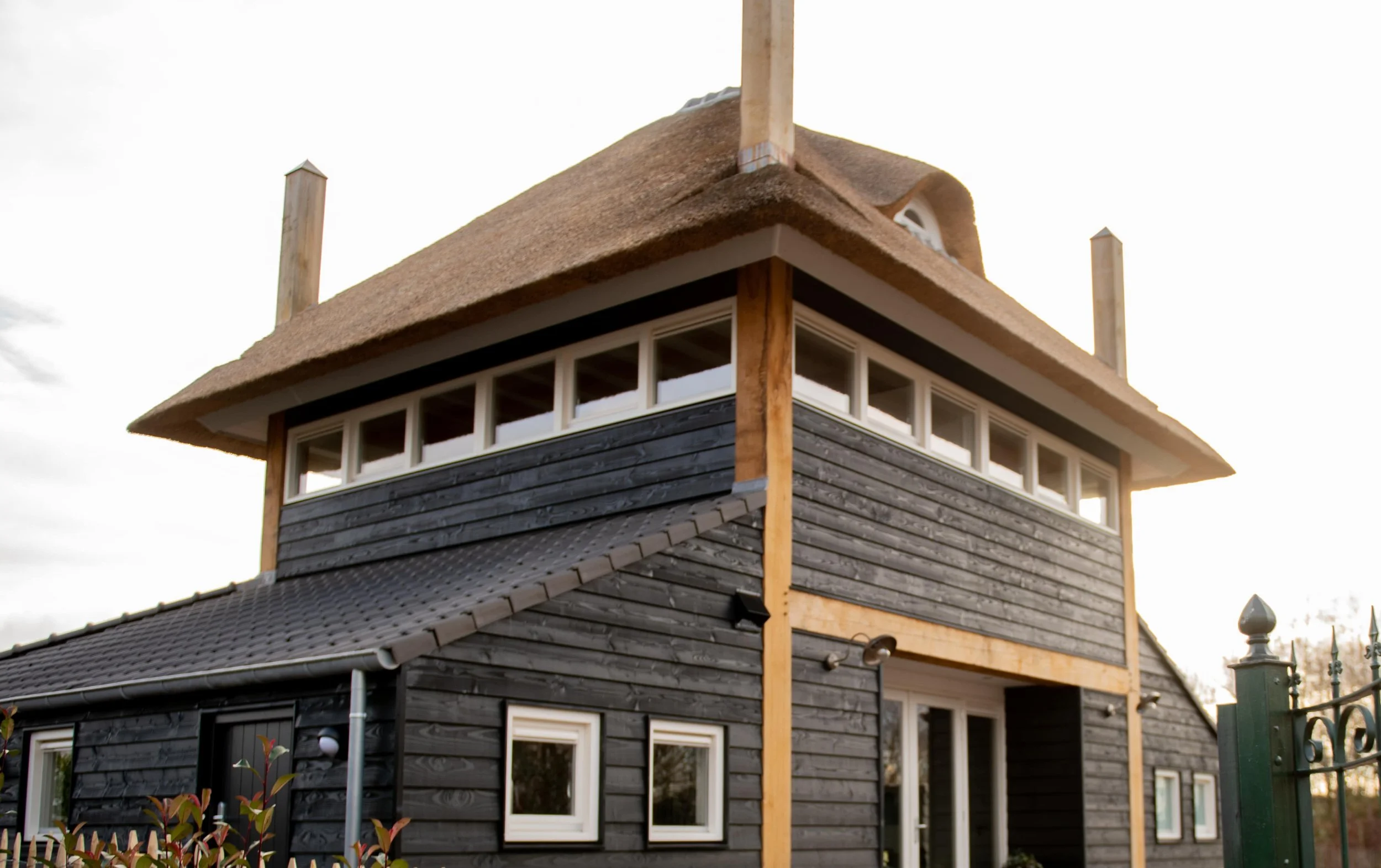 Moderne twee-onder-een-kap woning met zwarte houten gevel en rieten en dakpannen daken, omgeven door een tuin en een hek.