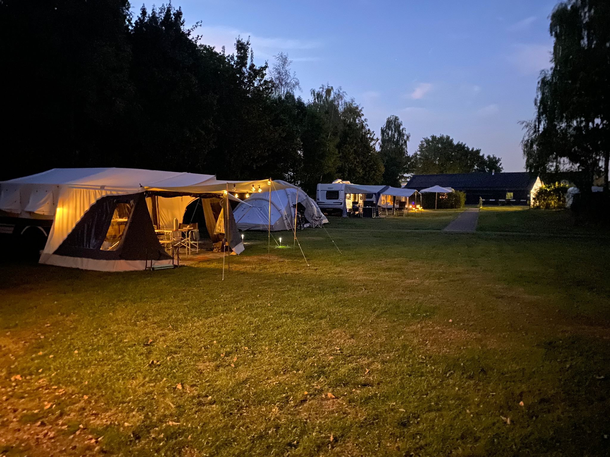 Een camping met tenten en caravans in de schemering, verlicht door zachte gele en witte lichten, omgeven door bomen en gras.