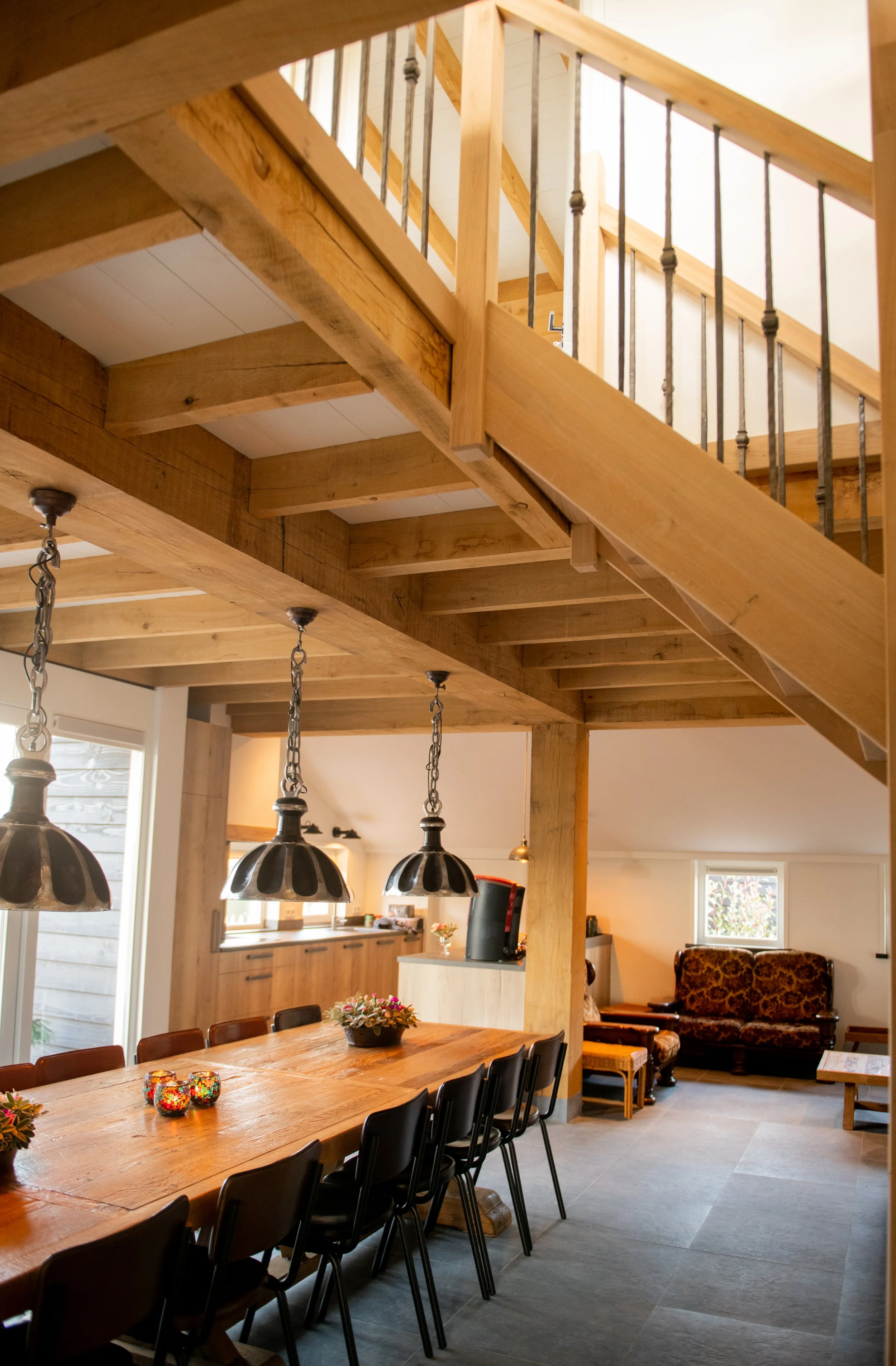 Interieur van een woonhuis met een eettafel, hanglampen, een bank, een venster en een open houten trap naar een bovenverdieping.