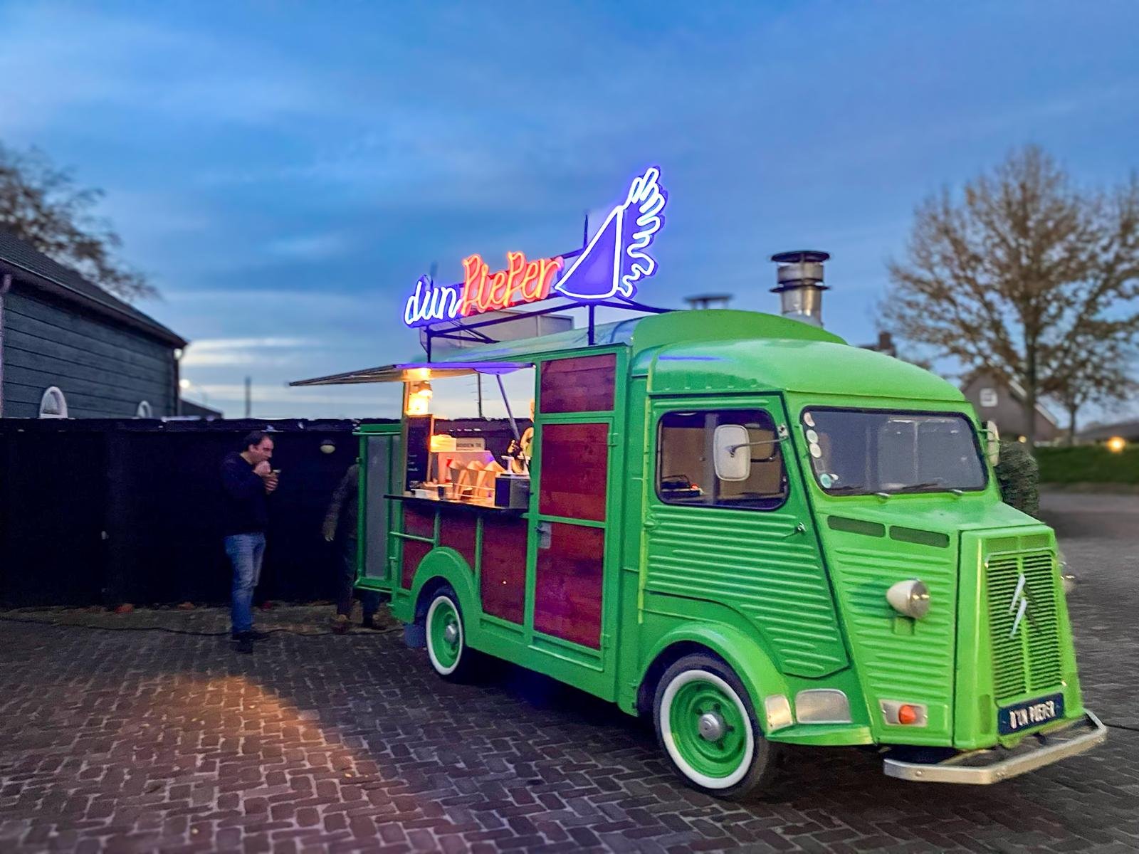 Groene vintage foodtruck met neonborden die 'duin Liefde' zegt, op een verlichte avond, met mensen eromheen.