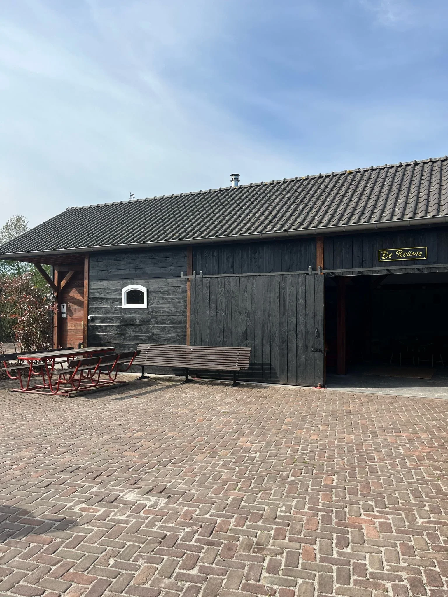 Een boerderij met een donkere houten gevel en een zadeldak met dakpannen. Er staan een bank, een tafel en enkele stoelen buiten, en er is een gedeeltelijke open schuifdeur.