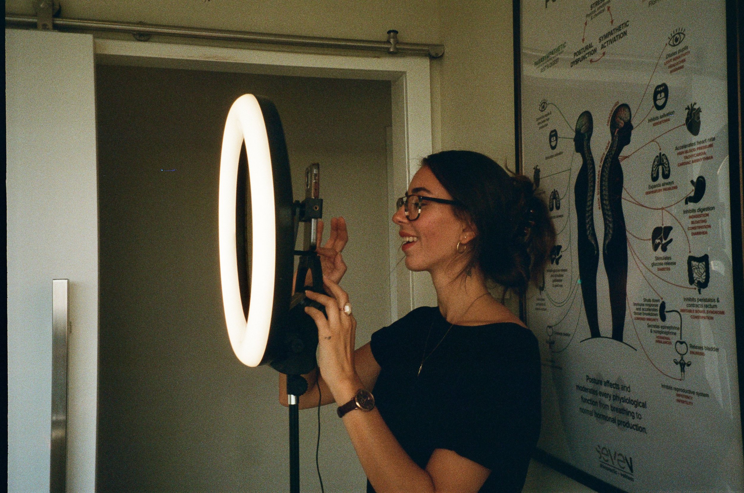 A woman with glasses and dark hair smiling while adjusting a camera with a ring light attached, standing next to a diagram of the human body's nervous system on the wall.