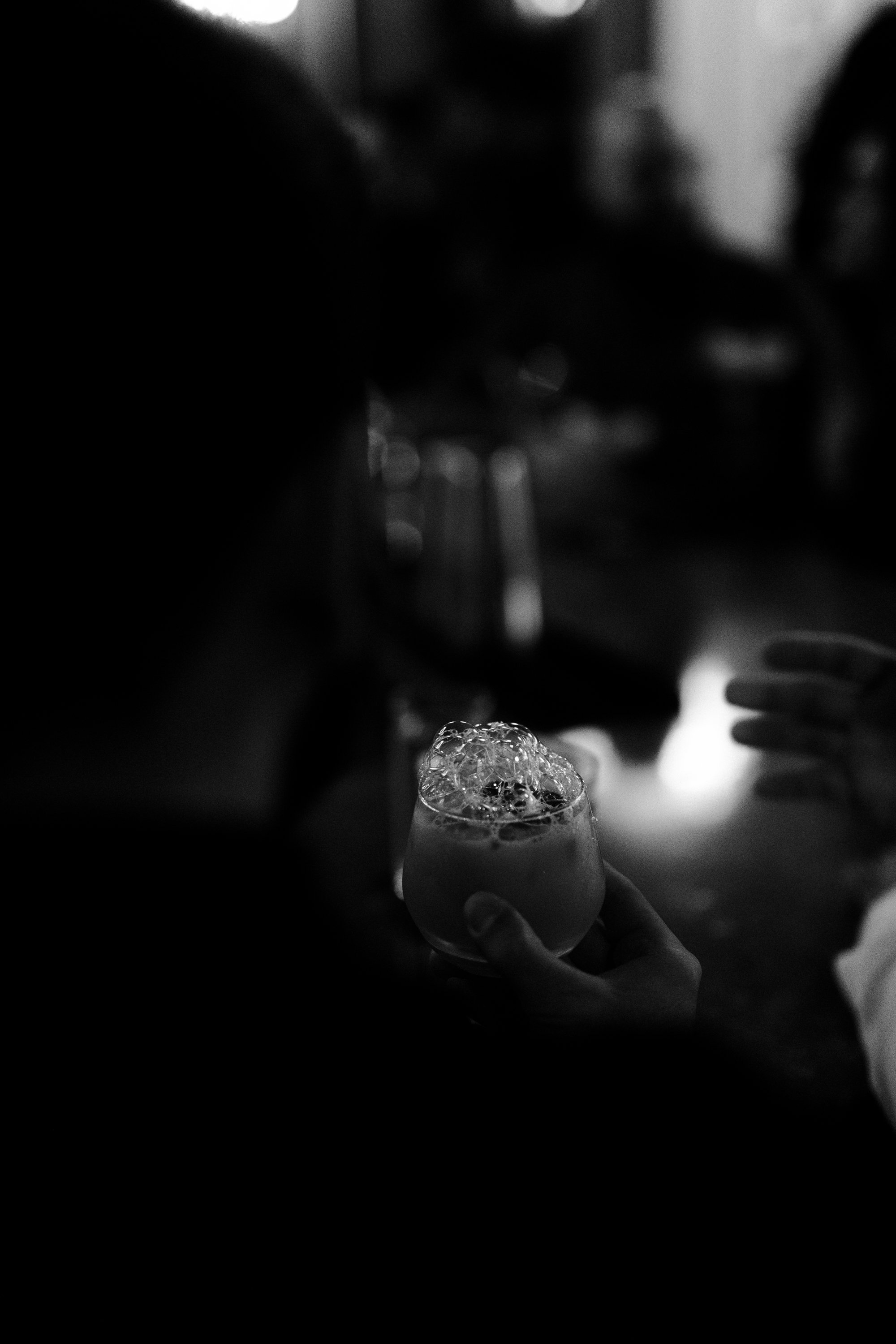 A person holding a glass of bubbly beverage in a dimly lit setting, with other people nearby, in black and white.