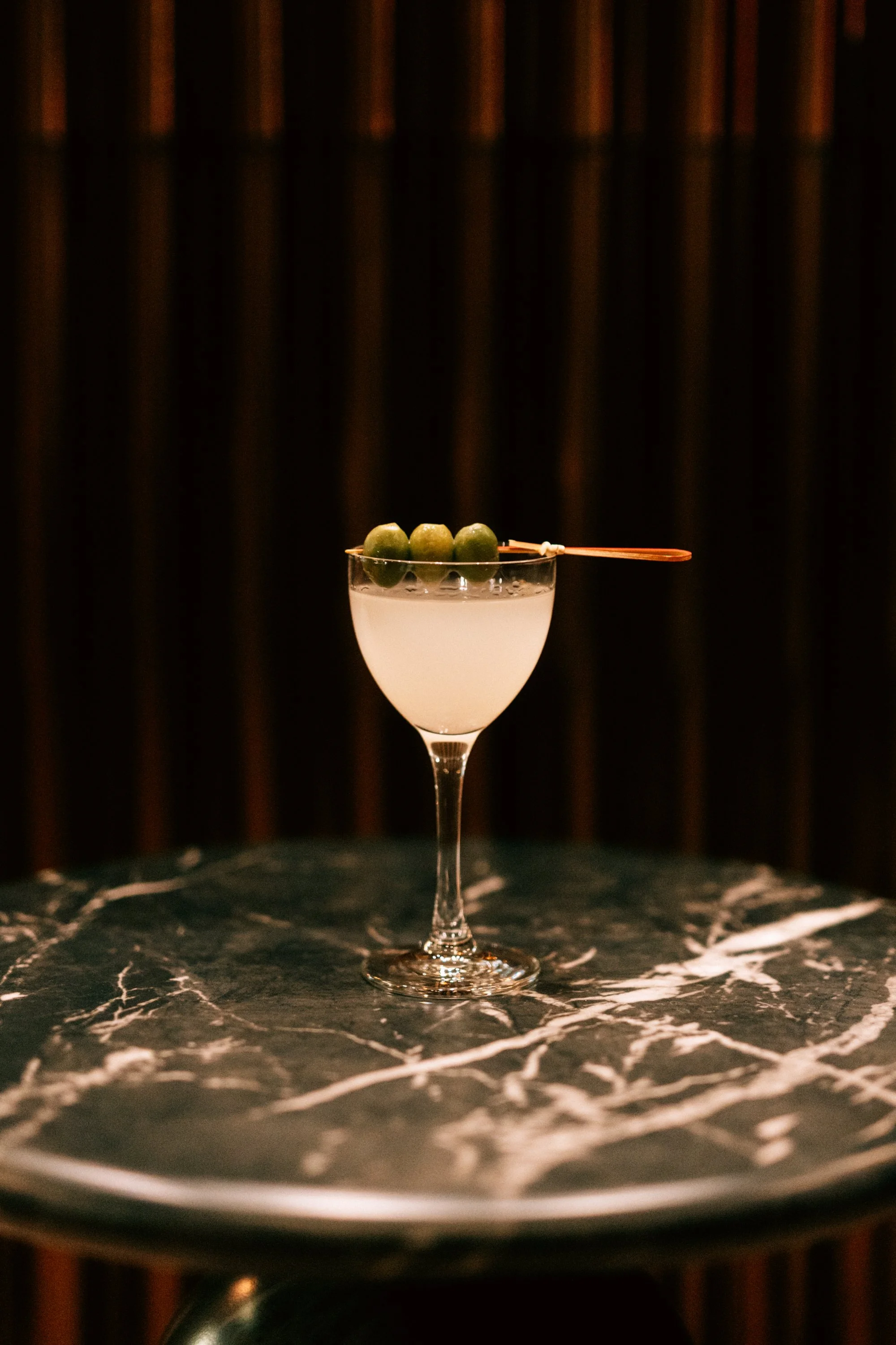 A cocktail glass with a light-colored drink, garnished with green olives on a skewer, resting on a dark marble table with white veining, and a dark background.
