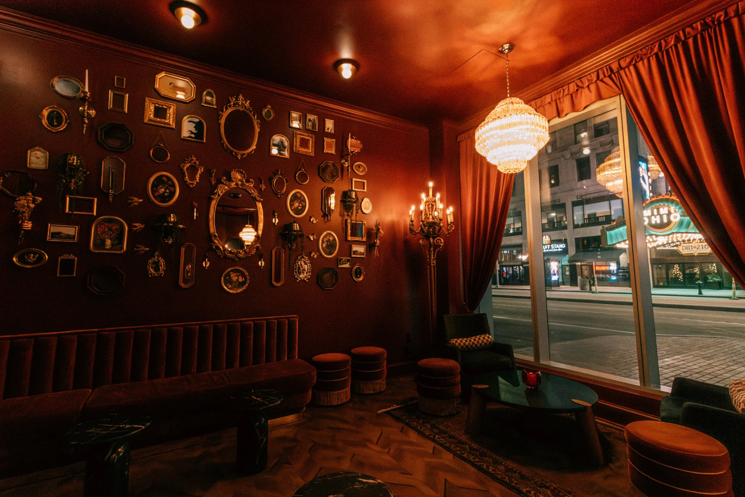 Interior of a vintage-style lounge with dark red walls, decorated with numerous small ornate mirrors and picture frames. There are velvet sofas, round tufted ottomans, a small coffee table, and a large window with heavy red curtains. Outside the wind