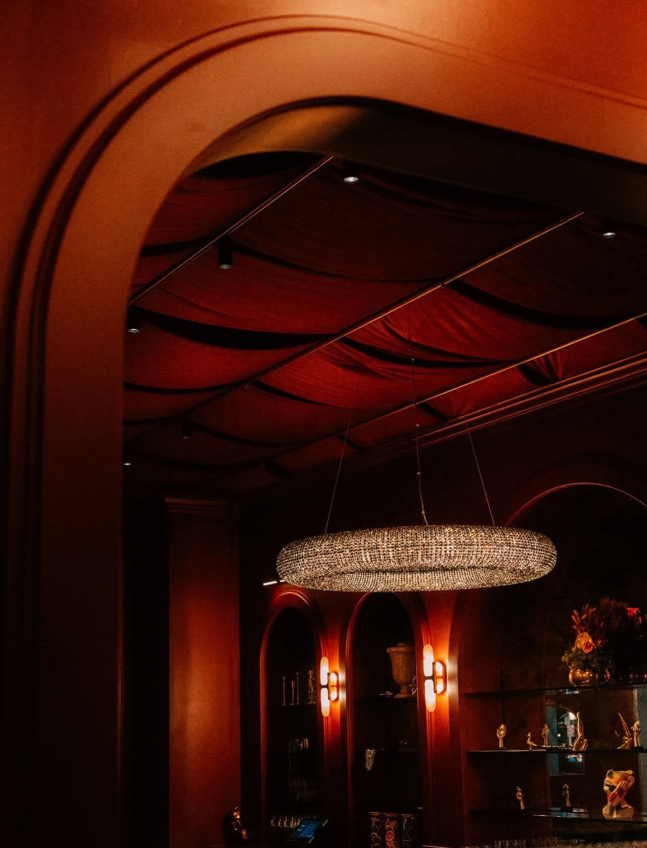 Interior view of a dimly lit room with a large, oval chandelier hanging from a curved ceiling with red fabric decor. There are arched shelves with decorative items on the dark wall with warm lighting.
