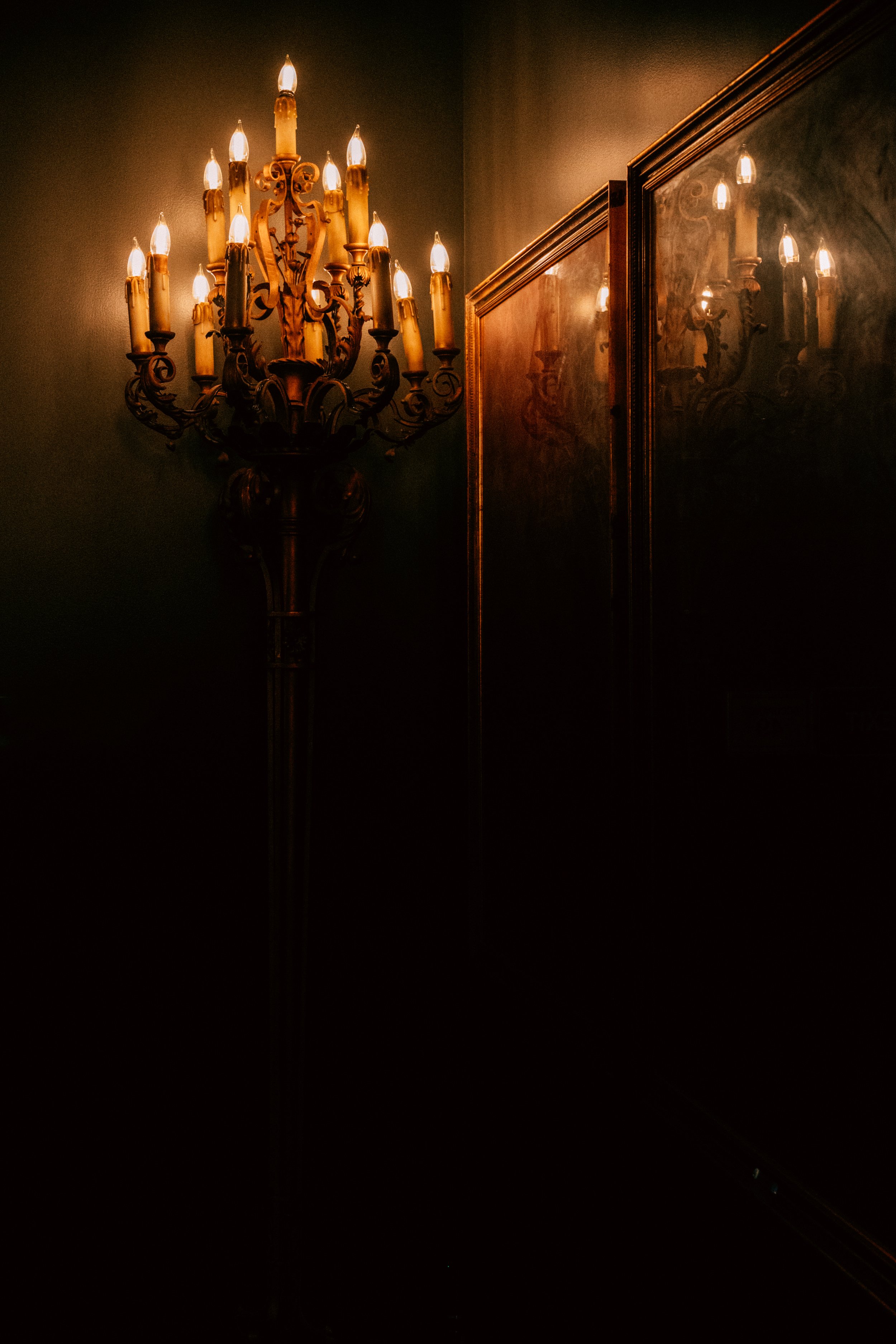 An ornate gold-colored chandelier with lit candles mounted on a wall, with two framed paintings or mirrors reflecting the chandelier's light.