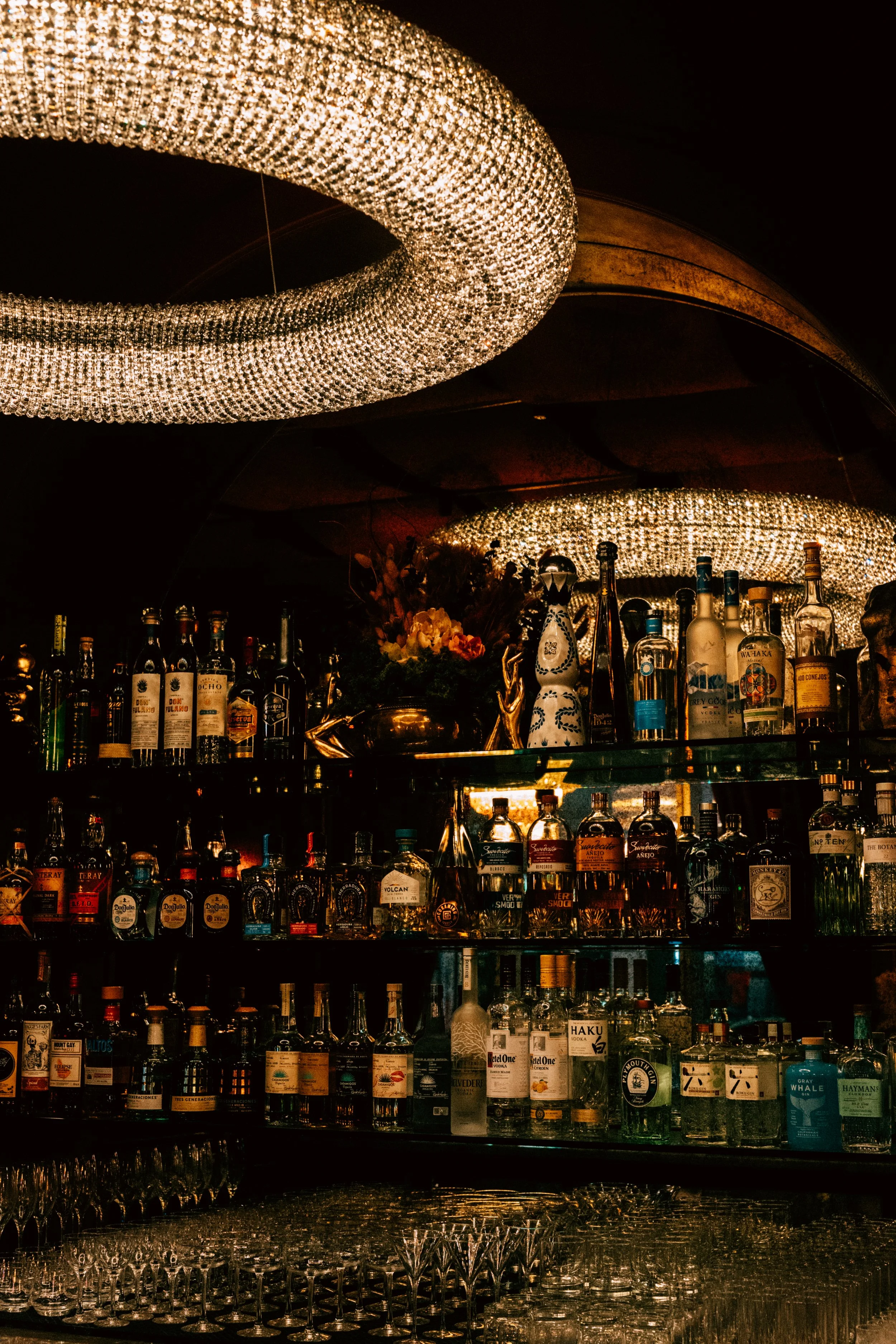 A well-stocked bar with various liquor bottles on shelves, illuminated by elegant chandeliers hanging from the ceiling.