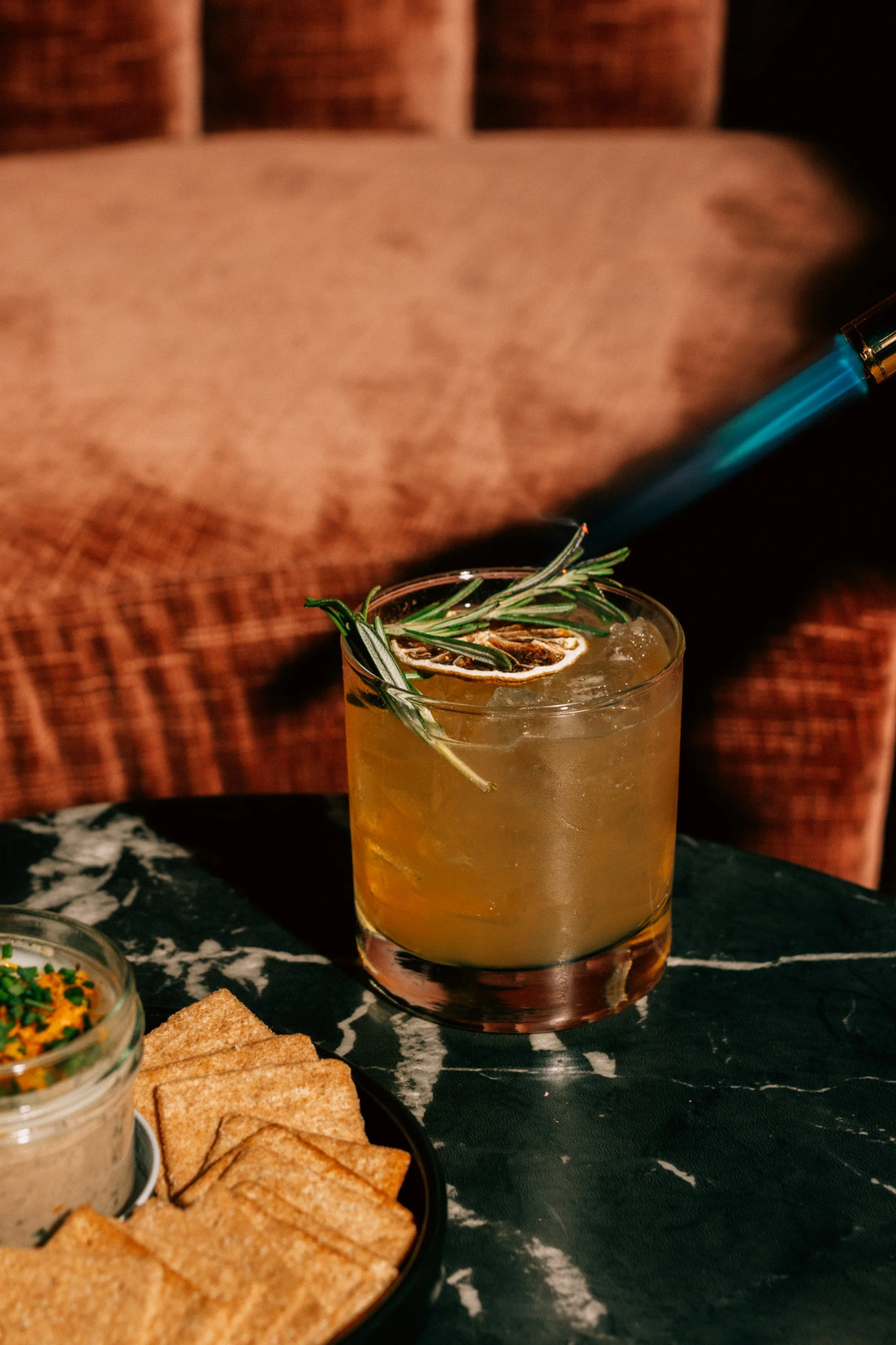 A cocktail drink garnished with rosemary and a dried lemon slice, served on a dark table with a bowl of crackers and a small bowl of dip nearby.