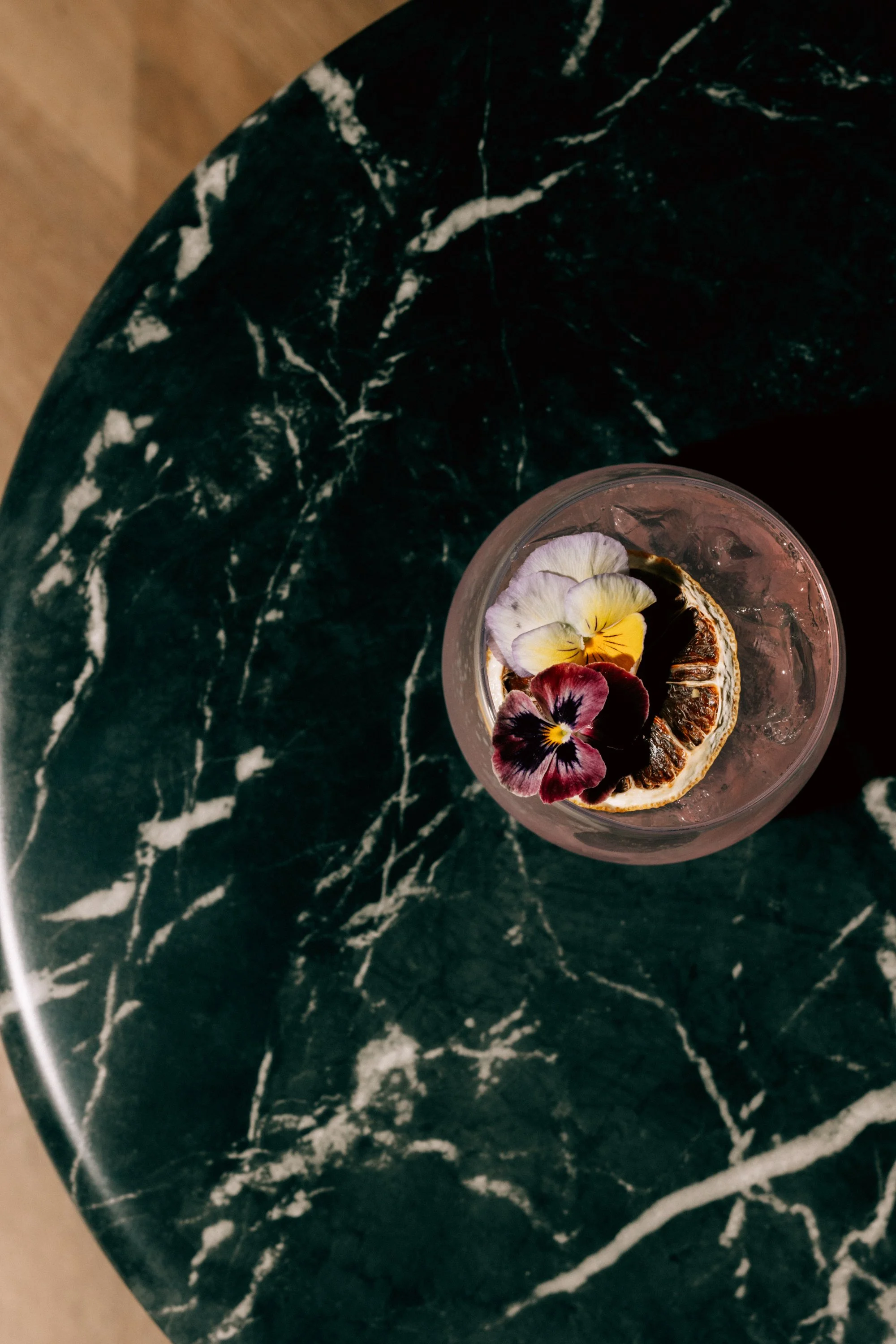 A clear glass of iced beverage garnished with dried orange slices and edible flowers on a dark green marble table.