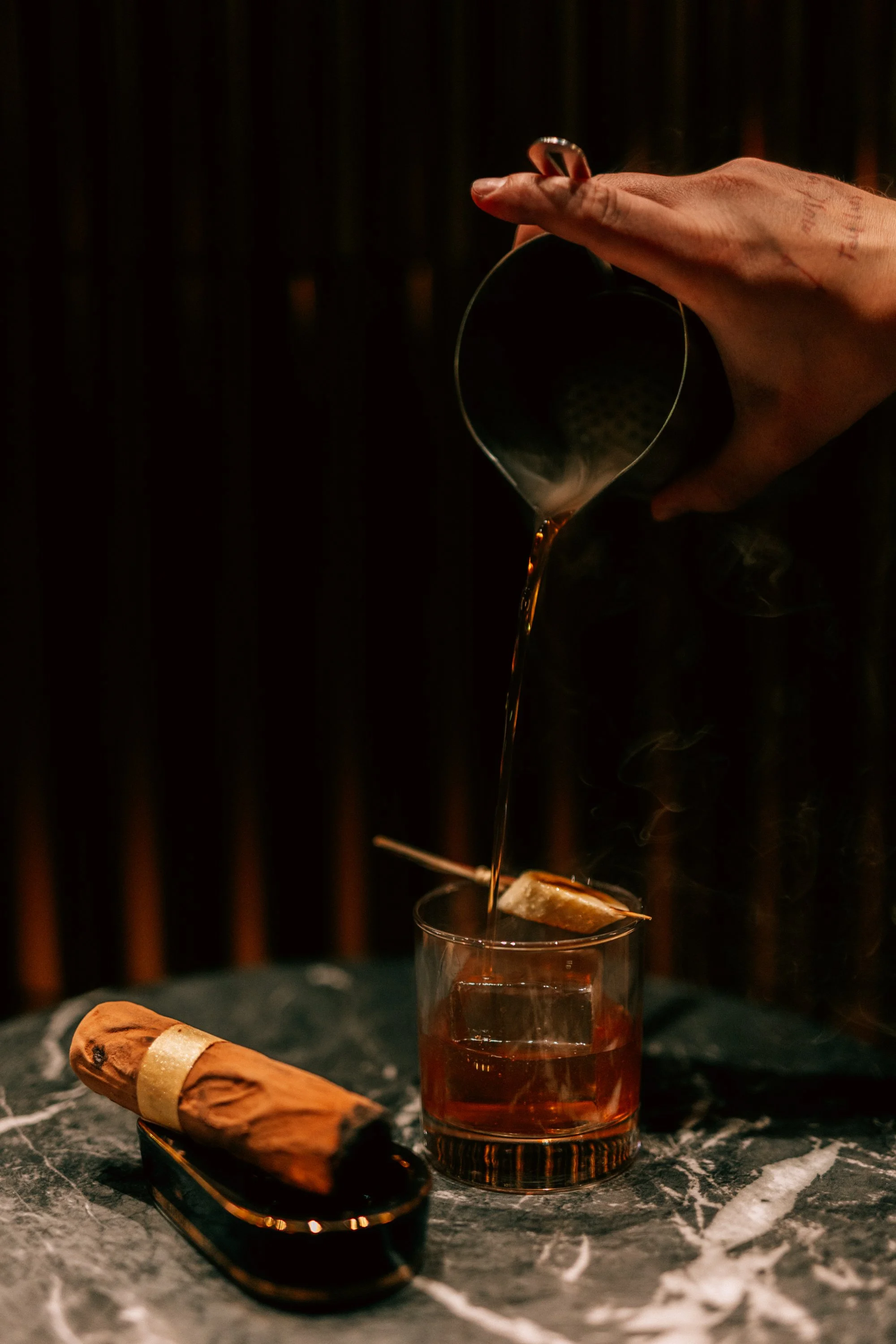 A hand pouring dark liquid into a glass with ice, next to a cigar on a black marble surface.