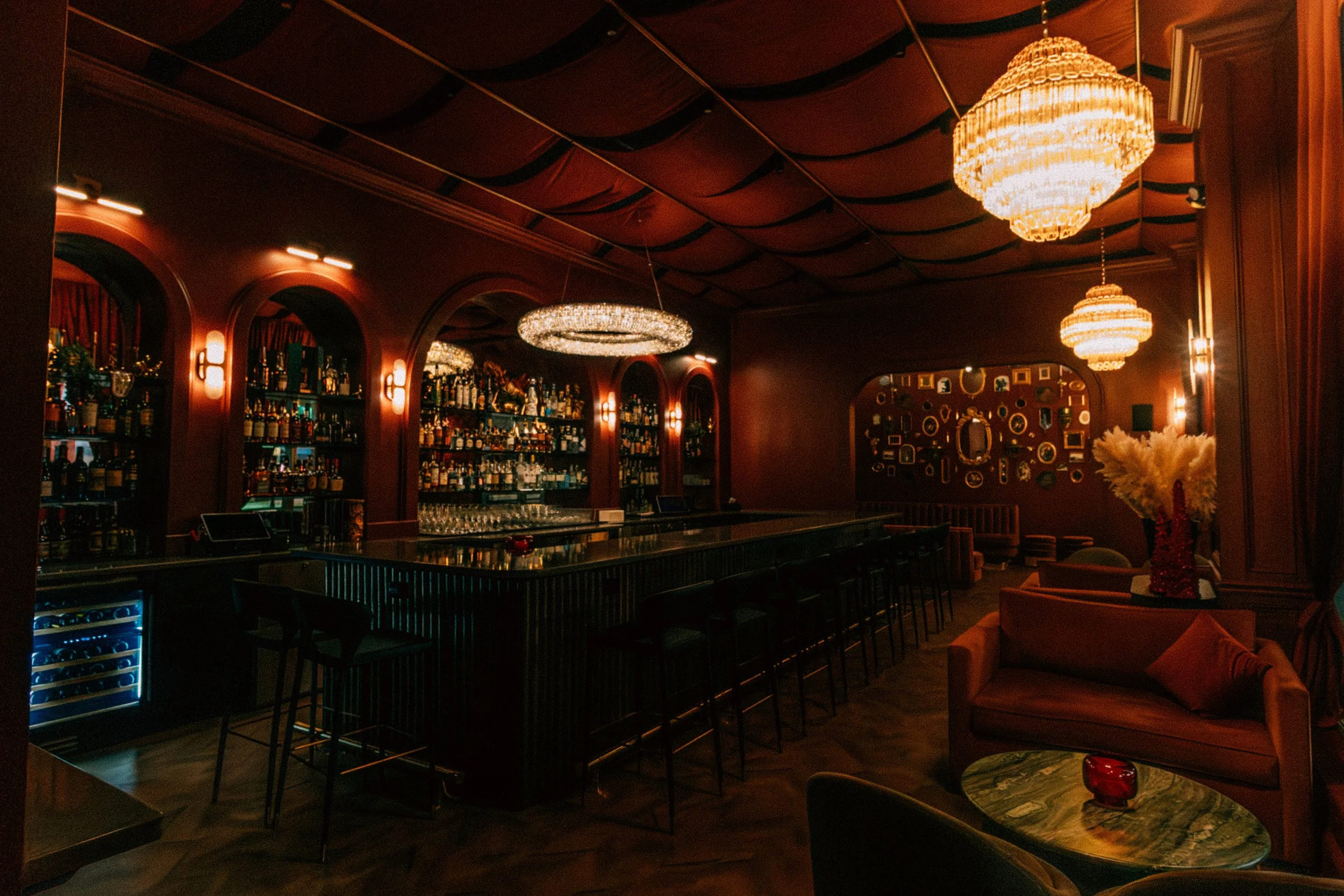 An elegant bar with a dark marble counter, surrounded by high stools and a backlit liquor shelf, warm lighting, chandeliers, a seating area with a sofa and armchair, and decorative wall art.