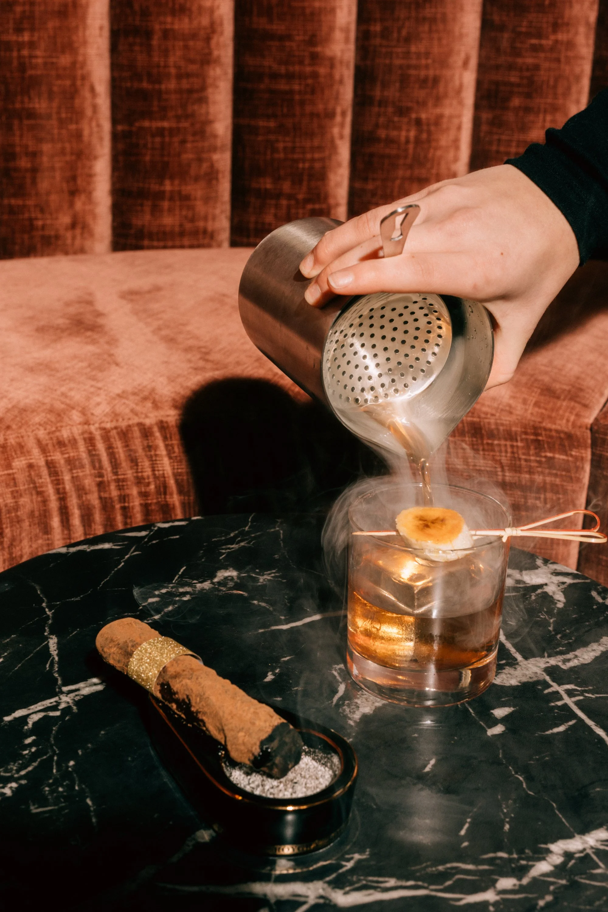 Person pouring hot liquid over a garnish in a glass, with a candied cigar on a black marble table.