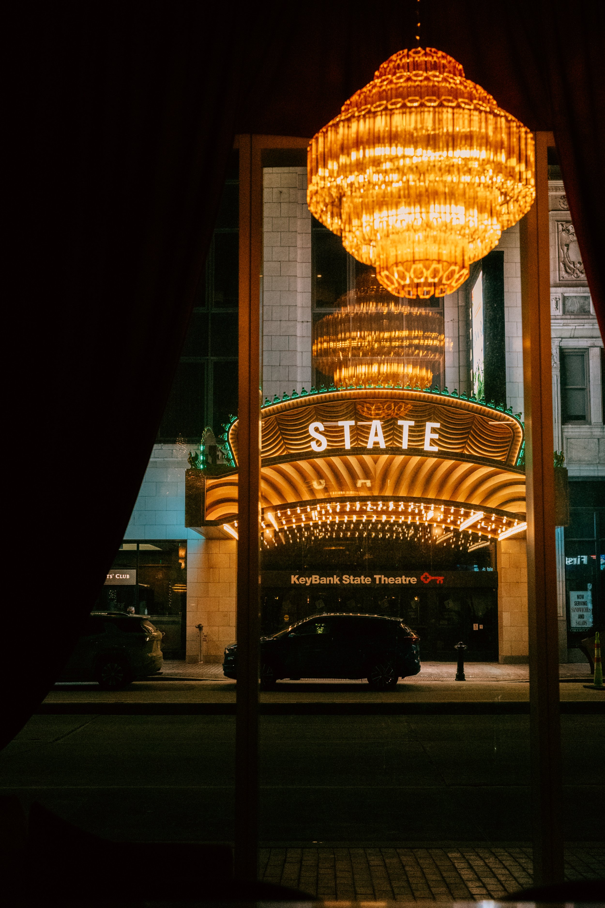 View of the KeyBank State Theatre entrance at night, with illuminated marquee and chandelier visible through a window, framed by dark curtains.