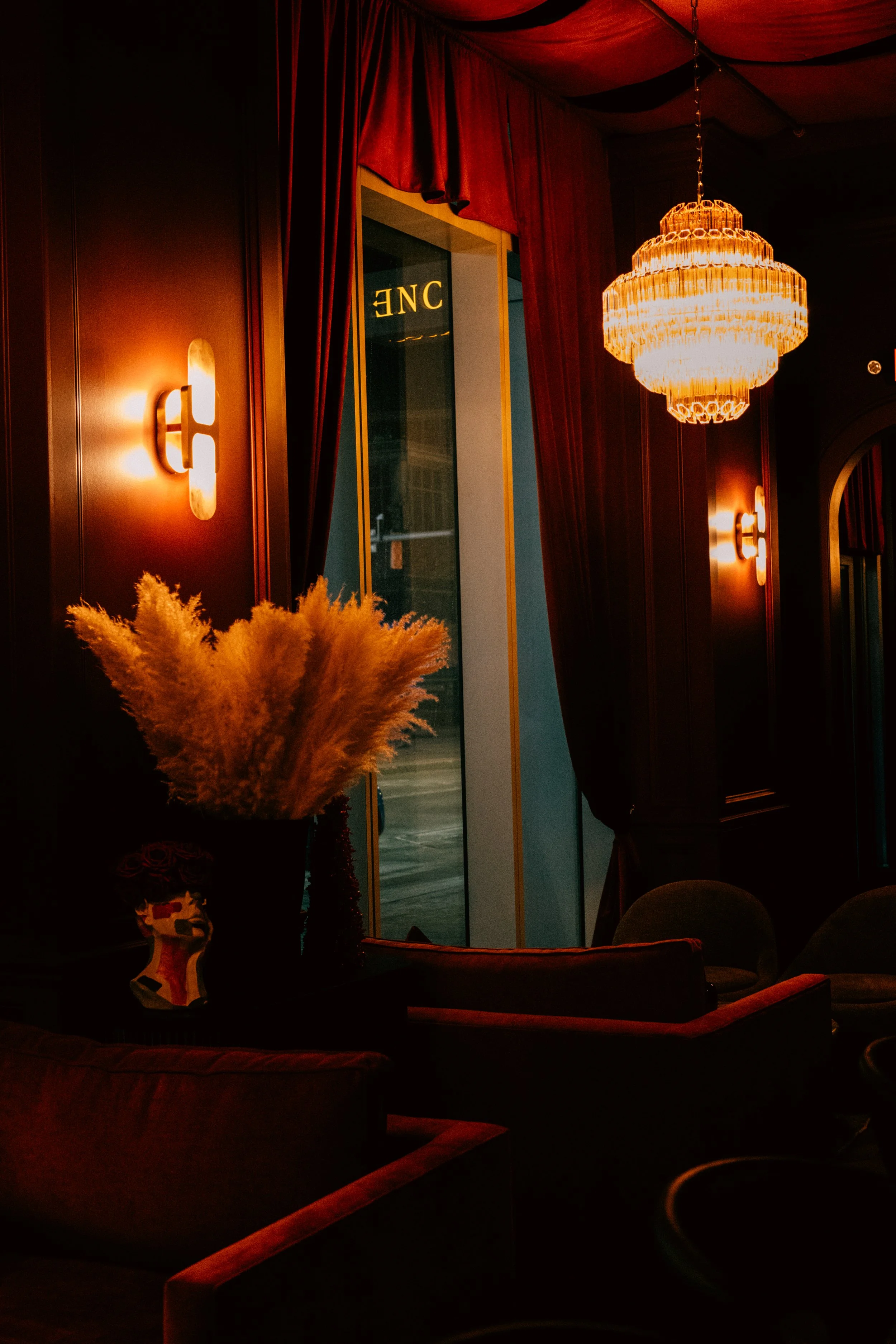 Dimly lit interior of a lounge or restaurant with a chandelier, wall sconces, velvet curtains, and a decorative vase with dried pampas grass.