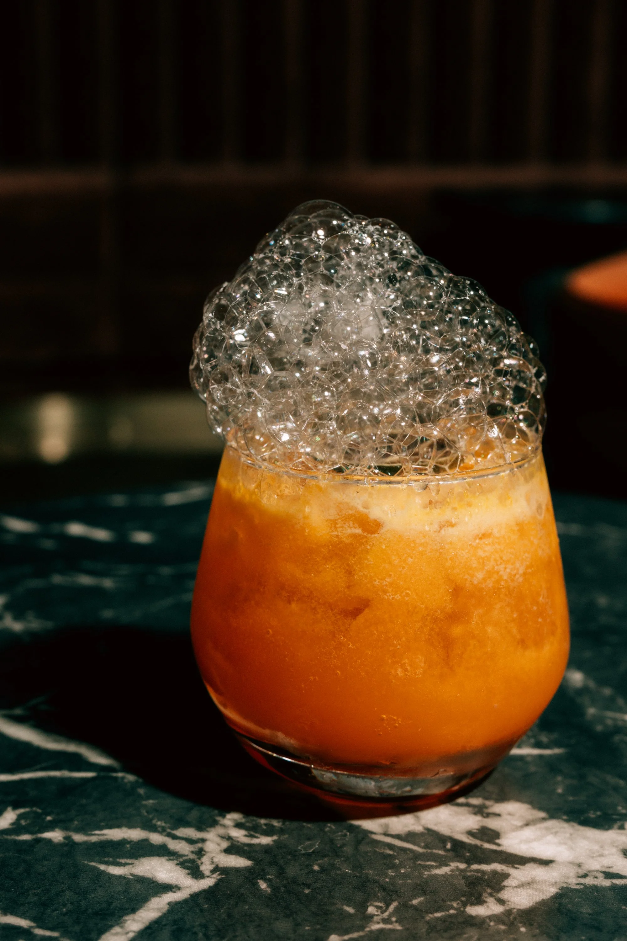 Close-up of an orange-colored drink in a rounded glass with foam and large bubbles on top, placed on a dark marble surface.