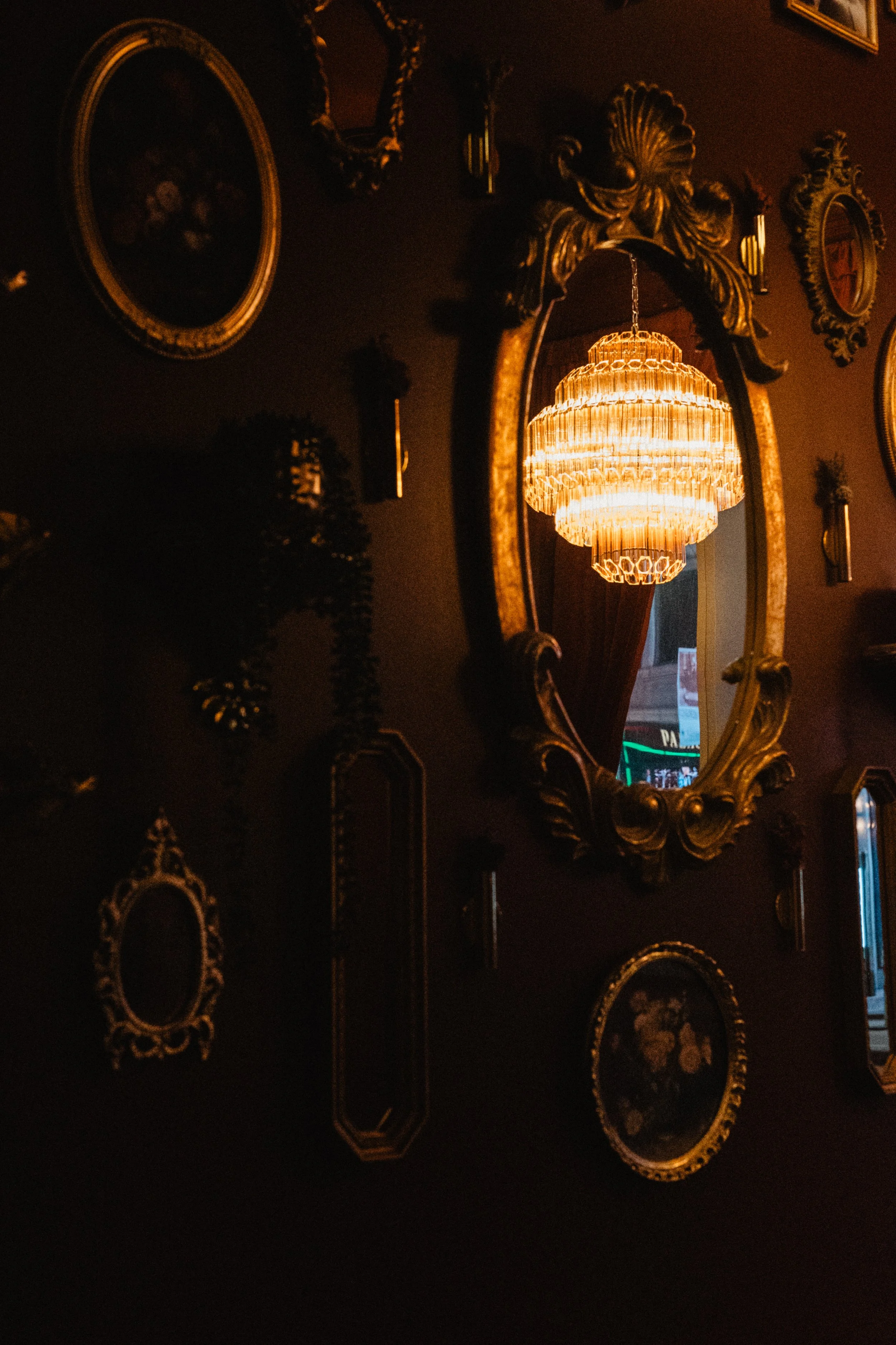 Decorative wall with multiple antique gold-framed mirrors and artwork, illuminated by a large, ornate chandelier reflected in an oval mirror.