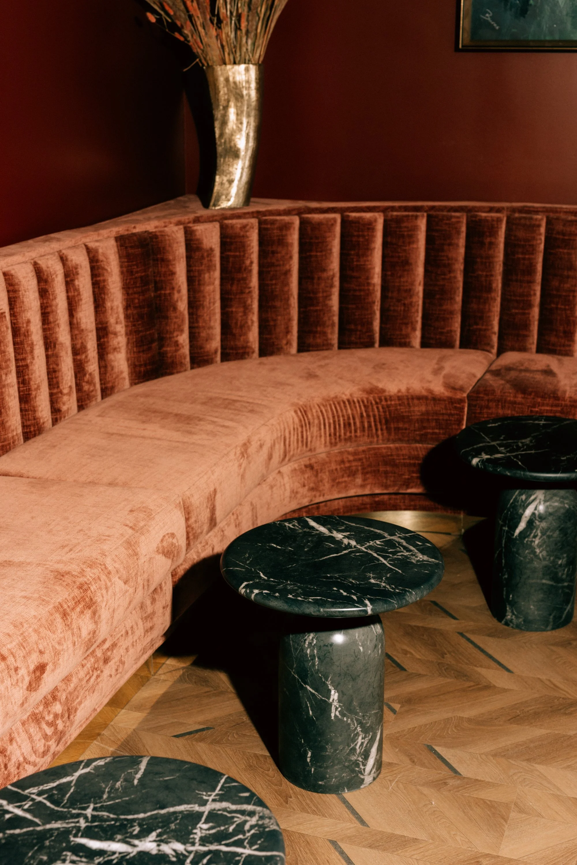 A blush pink velvet curved sofa with vertical channel tufting, dark green marble round side tables with white veining, a tall silver vase with dried branches, and a framed artwork on a dark red wall.