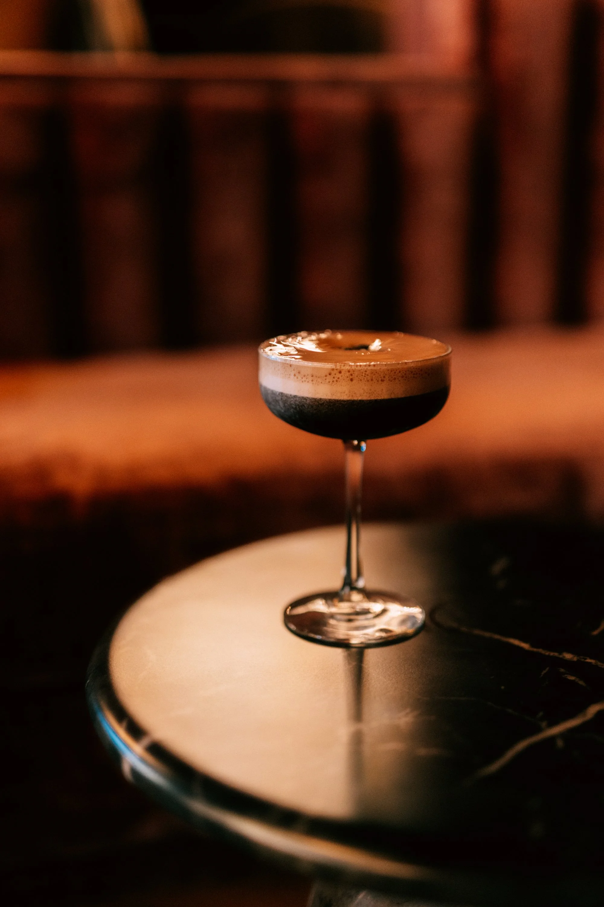 Close-up of a cocktail in a champagne coupe glass on a reflective black surface with a blurred wooden background.