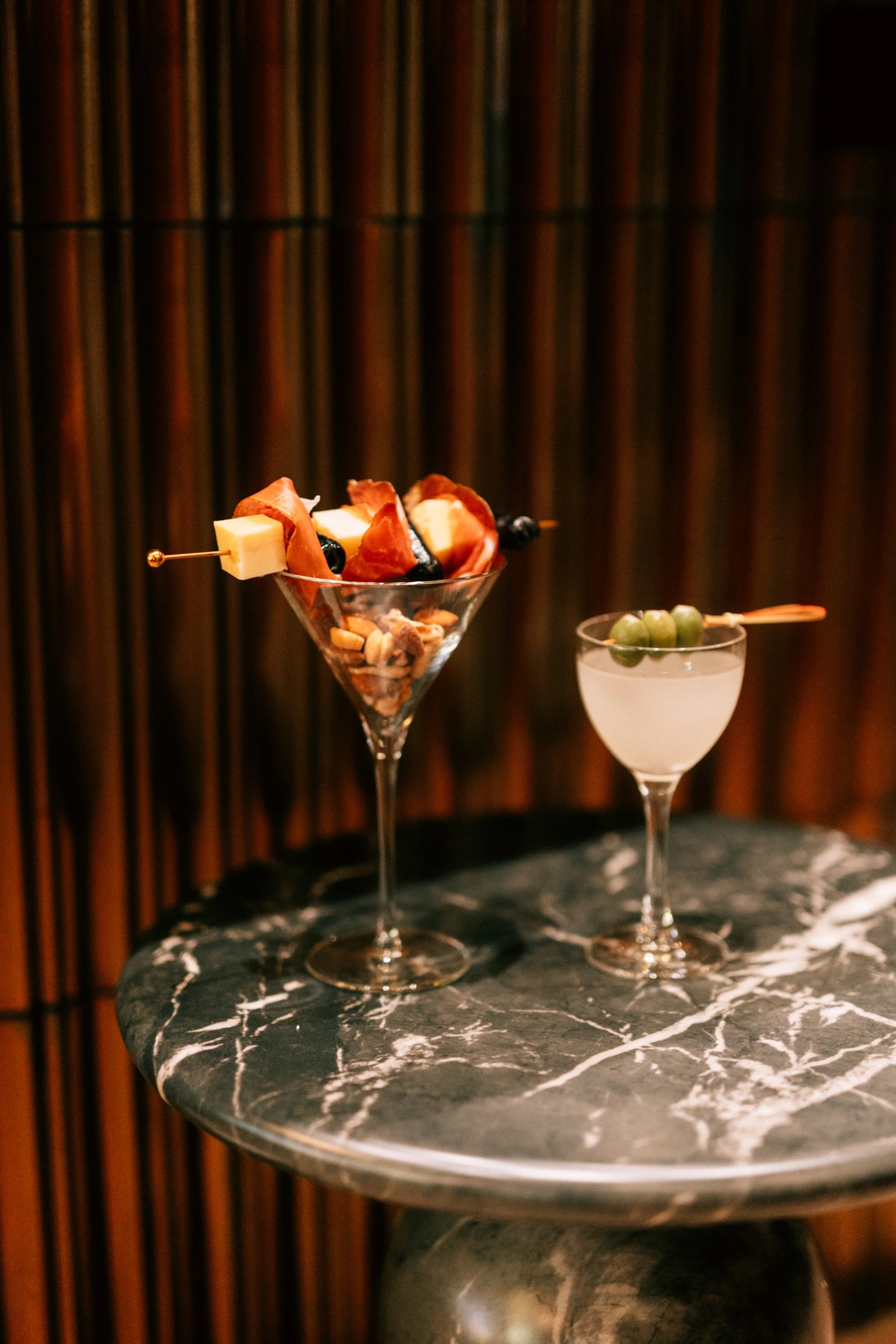 Two cocktails on a black marble table, an martini glass with a savory skewer and a smaller glass with green olives and a toothpick, wood panel background.