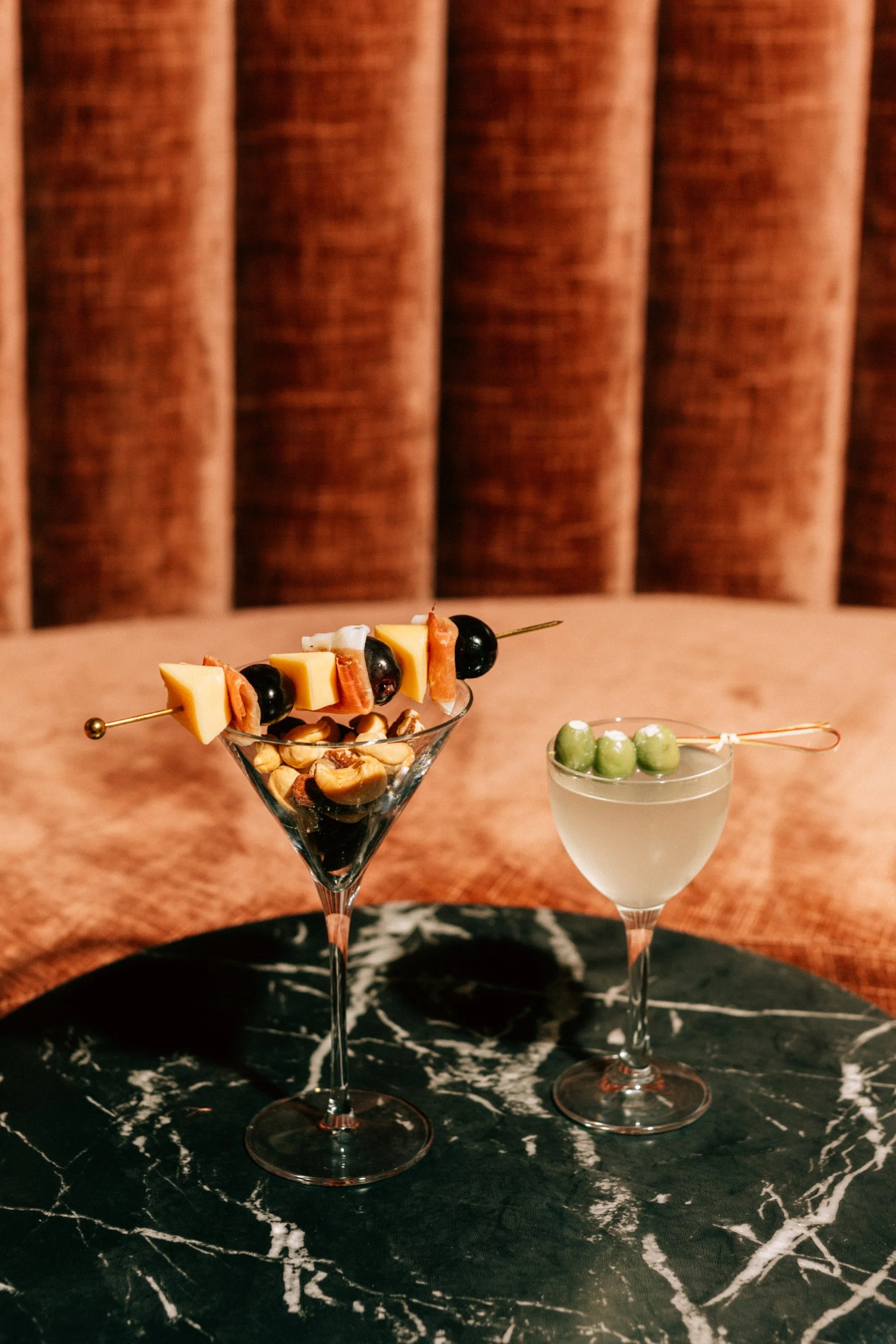 Two cocktail glasses on a black marble table, one with cheese, olives, nuts, and a skewer, the other with green olives and a clear liquid, against a textured brown background.