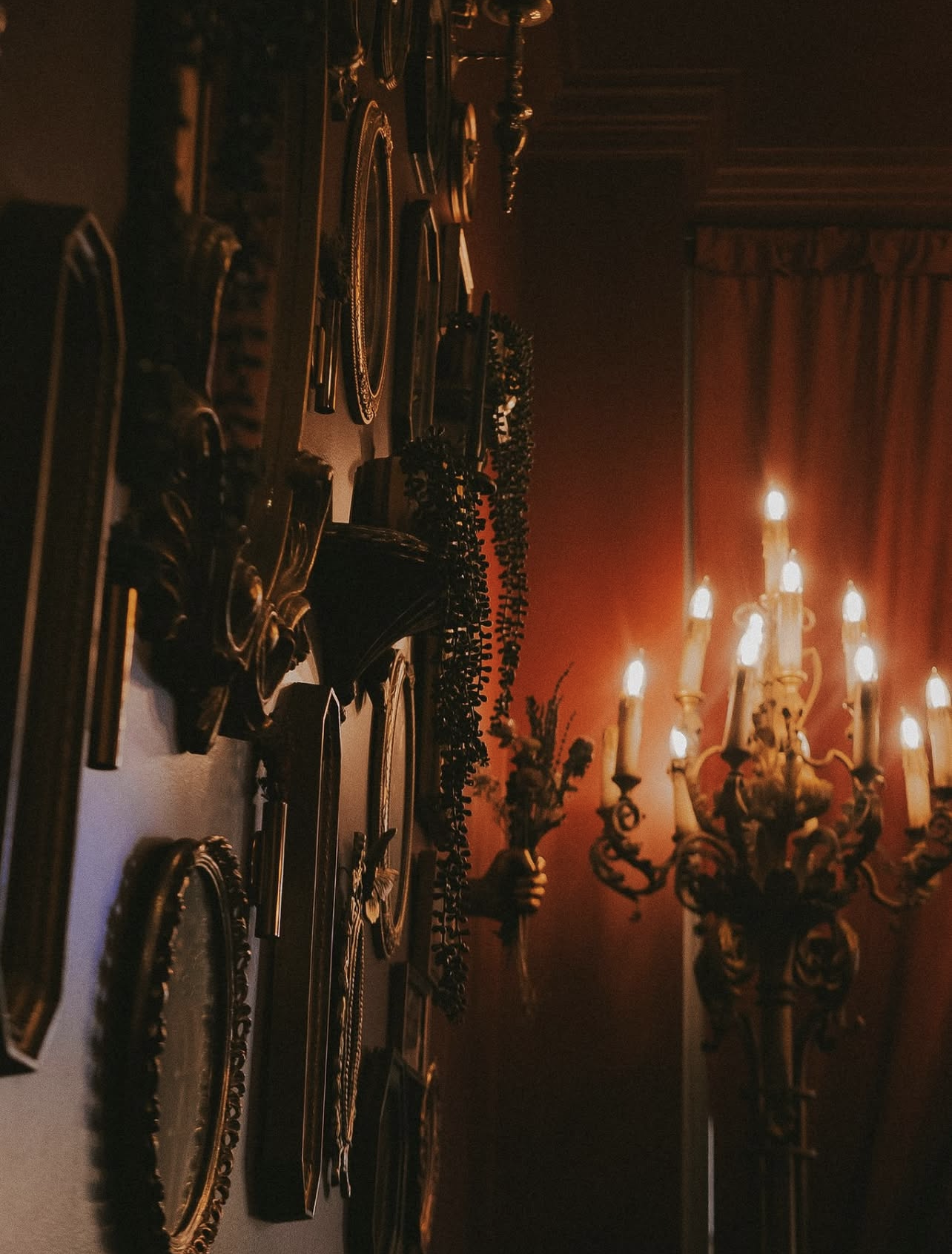 A dimly lit room with antique photo frames on a wall and a vintage chandelier with lit candle-shaped bulbs.
