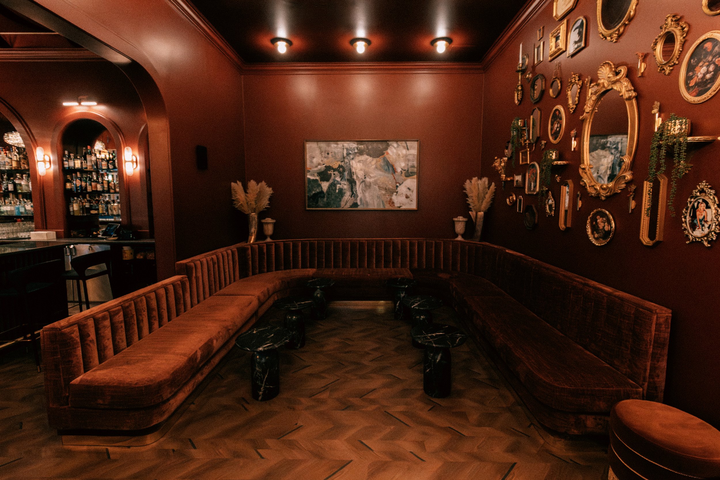 A cozy, dimly lit lounge with a large, curved velvet sofa, a gallery wall with various picture frames, and a bar area in the background with shelves of alcohol bottles.