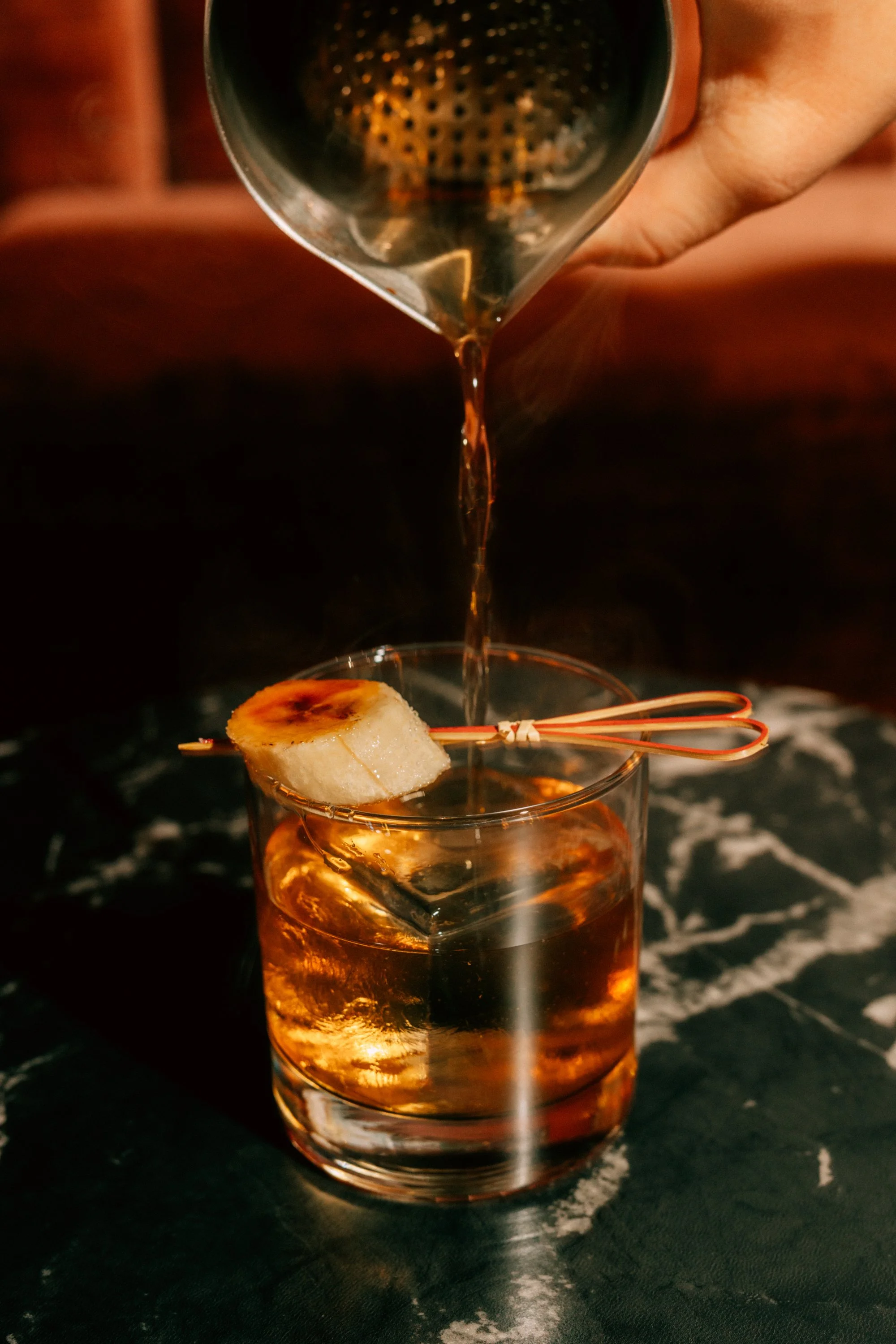 A hand pouring a drink over a large ice cube with a grilled banana slice and a cocktail pick in a short glass.